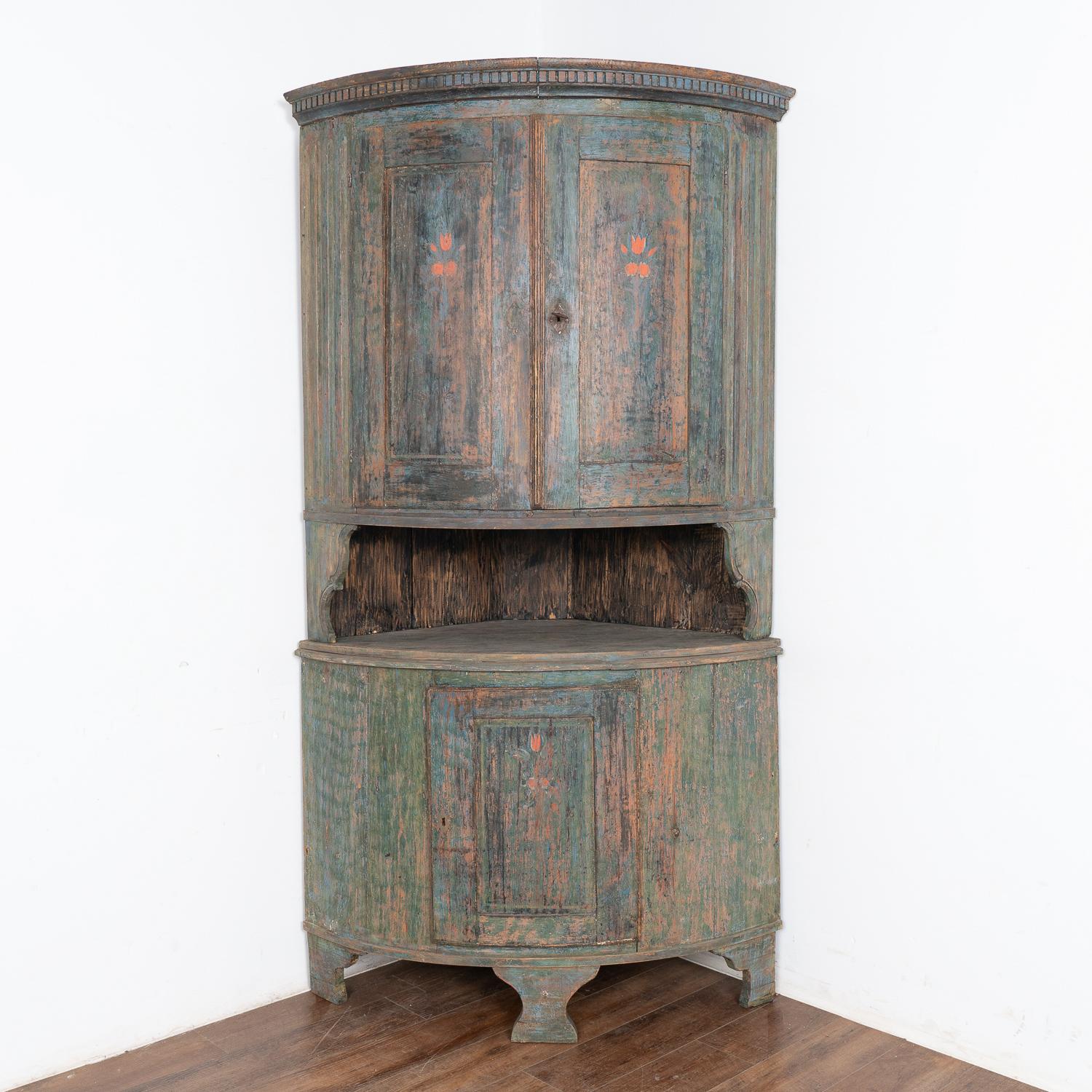 This tall bow front pine corner cabinet still maintains its original hand-painted finish with simple floral details remaining along the panels and topped with traditional dentil molding.
The original blue/green/teal painted finish has a soft, worn
