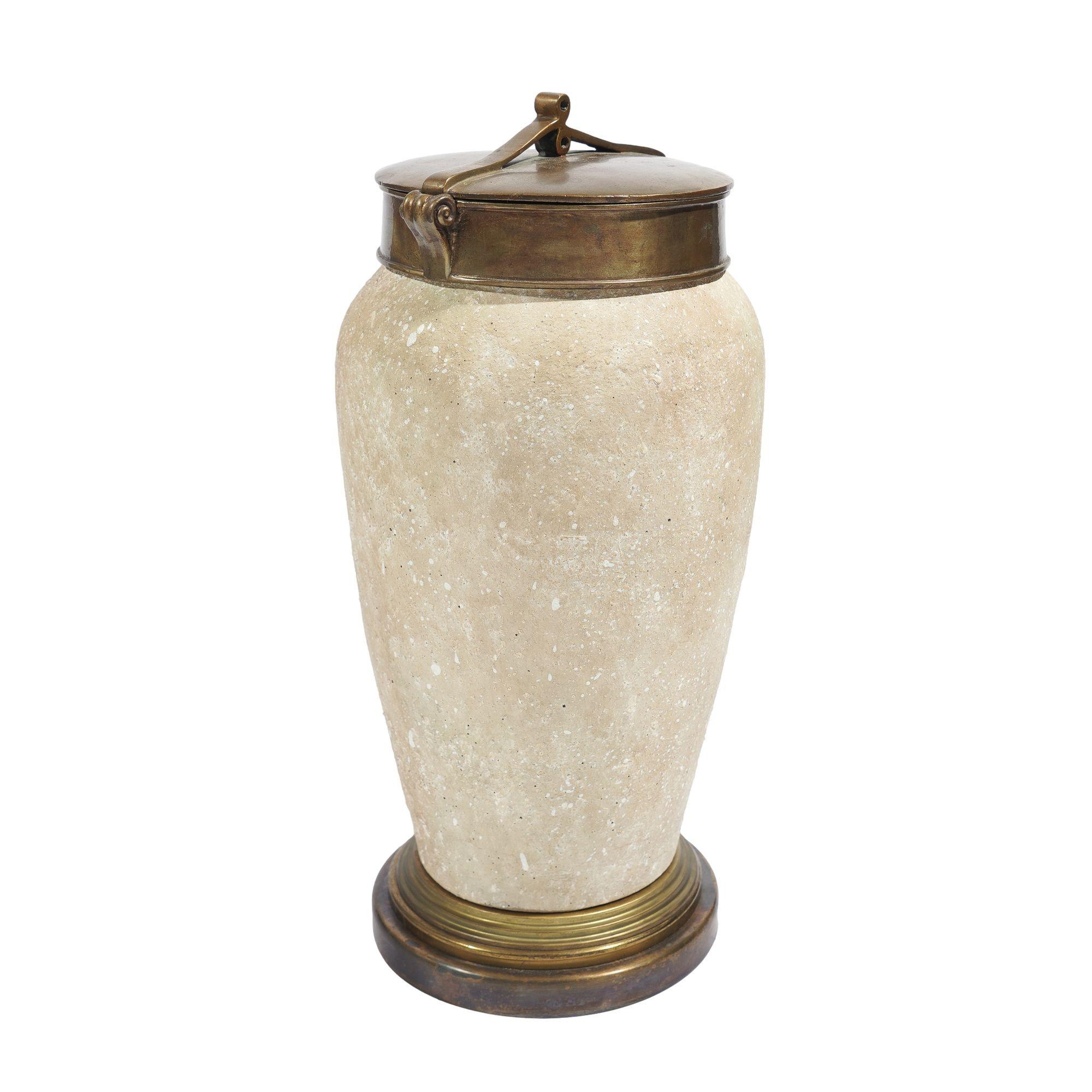A high shouldered art pottery urn with a tapered body in travertine tan with a textured surface. The urn is fitted with a wide, oxidized cast brass rim with flanking volutes and a lid with a strap work handle/finial. The brass base has a two stepped