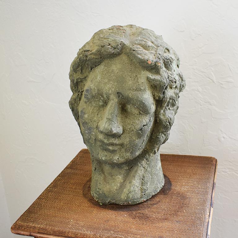 Concrete or stone bust of a man (or woman). The subject has curly wavy hair and is shown from head to neck. A beautiful piece to add a special conversation piece to any room. We love the idea of styling him with a hat.