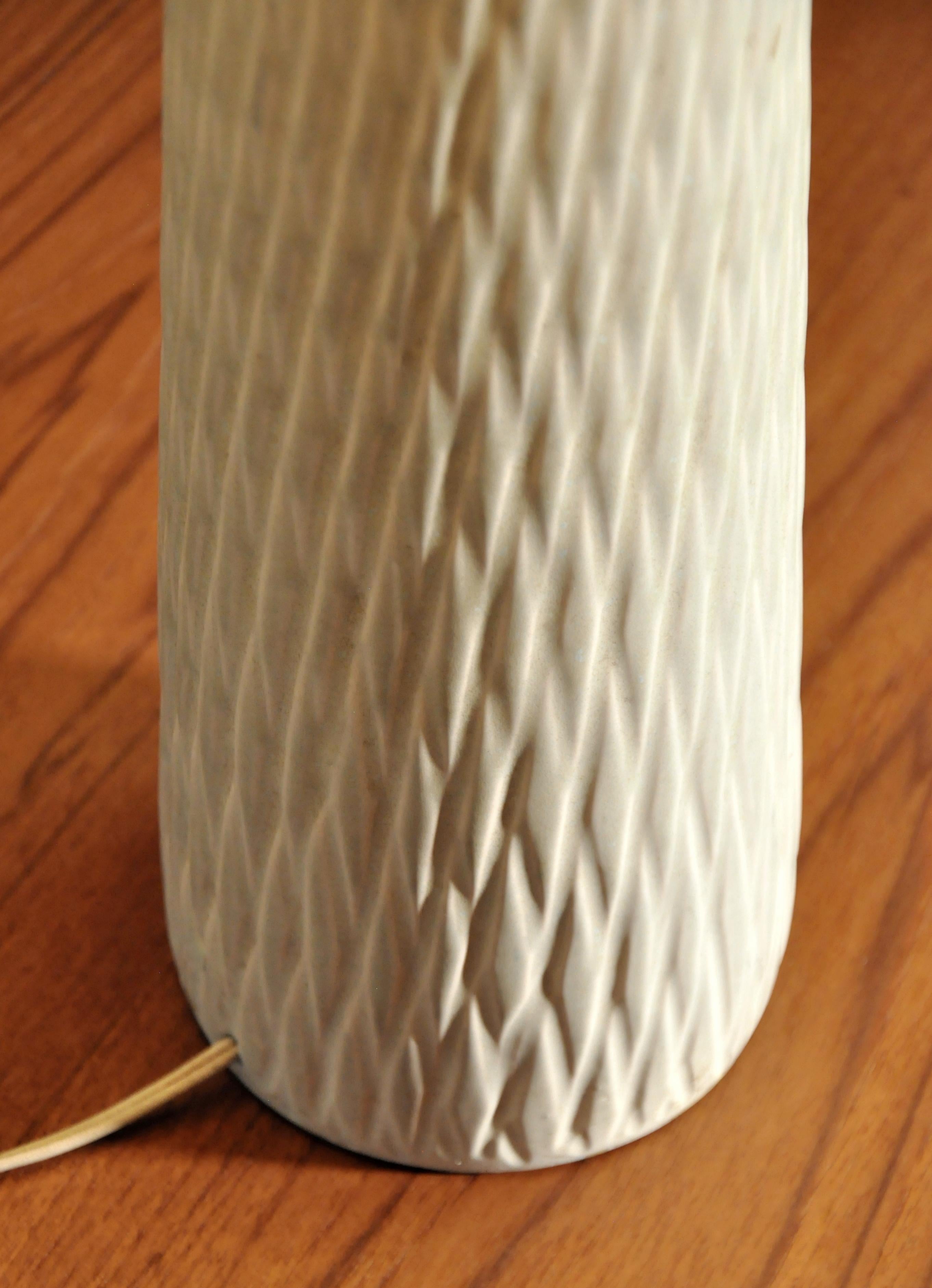 An off-white ceramic table lamp, dating from the late 1950s, attributed to Lotte & Gunnar Bostlund. The tall, vintage earth tone lamp features a creamy ivory colored body with a hand carved beehive pattern and a beige jute shade. Works well with