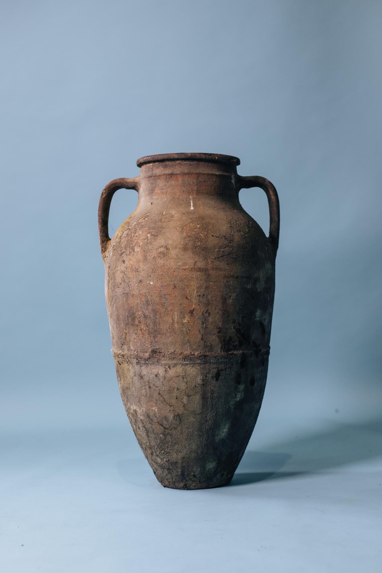 Questo vaso alto a doppio manico in terracotta è un bell'esempio di ceramica mediterranea dell'inizio del XX secolo: pratico nell'origine ma scultoreo nella forma. Realizzato a mano con argilla grossolana, un tempo serviva per conservare l'olio