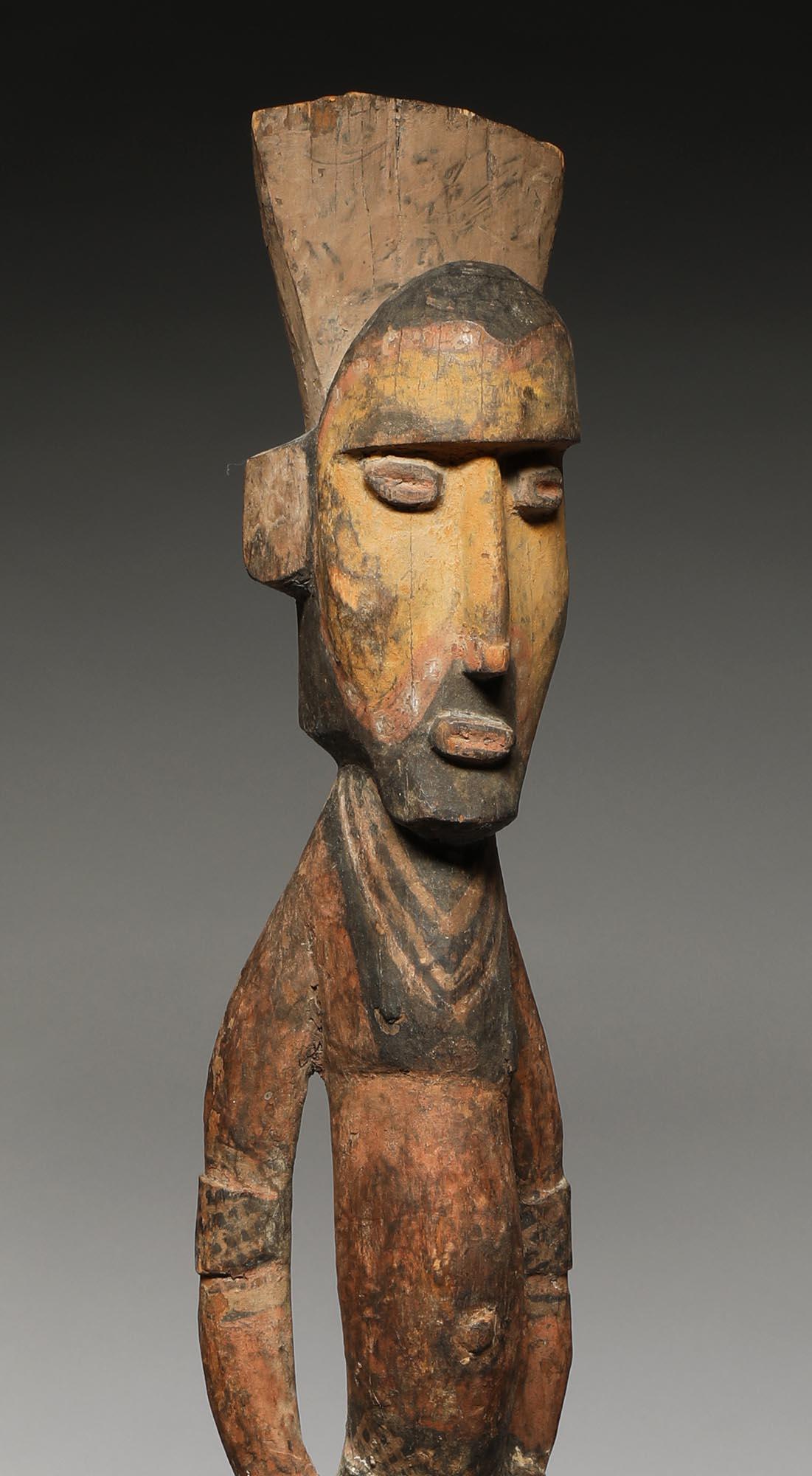A tall thin standing figure carved of hard wood, with hands on hips, wearing armbands, with remains of yellow pigments on face, black beard and top of head, traces of red on body. Classic Maprik figure with strong expression, on custom metal base 39