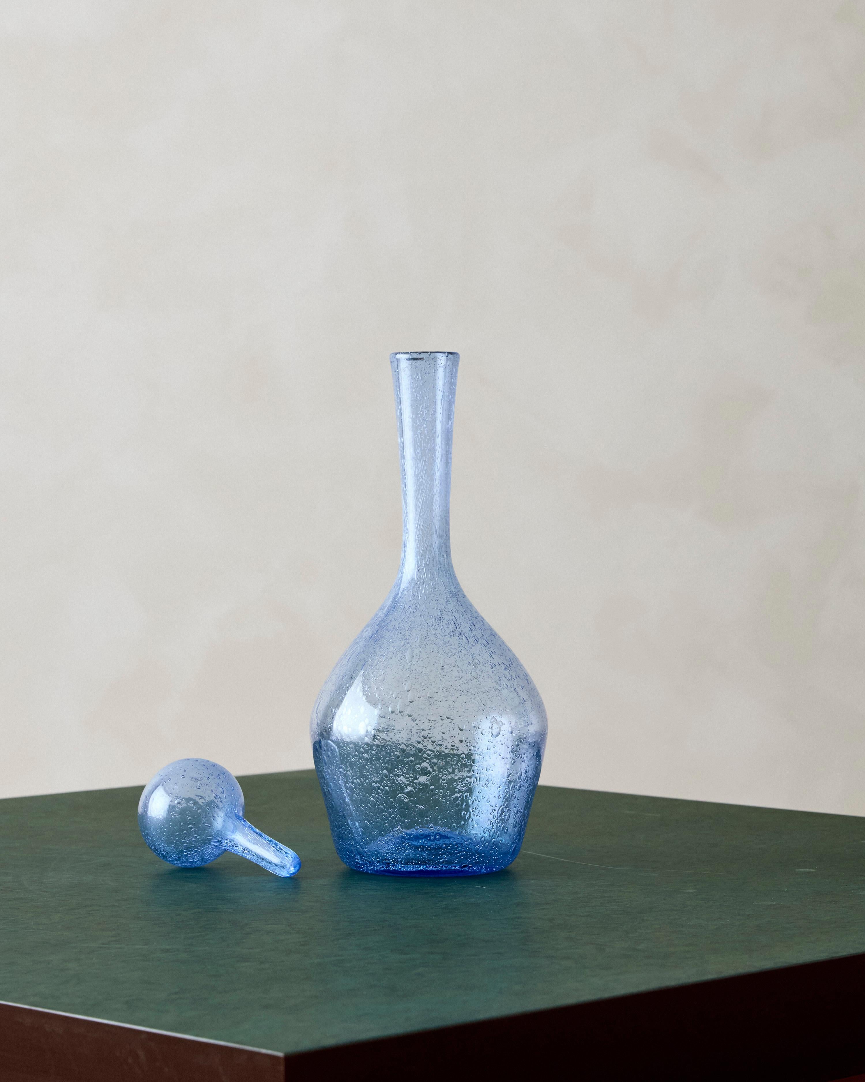 Caraffa francese soffiata a mano di colore blu traslucido, caratterizzata da un collo lungo con bolle che si estendono per tutto il vetro. Ha una base quadrata con inserti concavi rotondi su ogni lato ed è rifinito con il caratteristico tappo