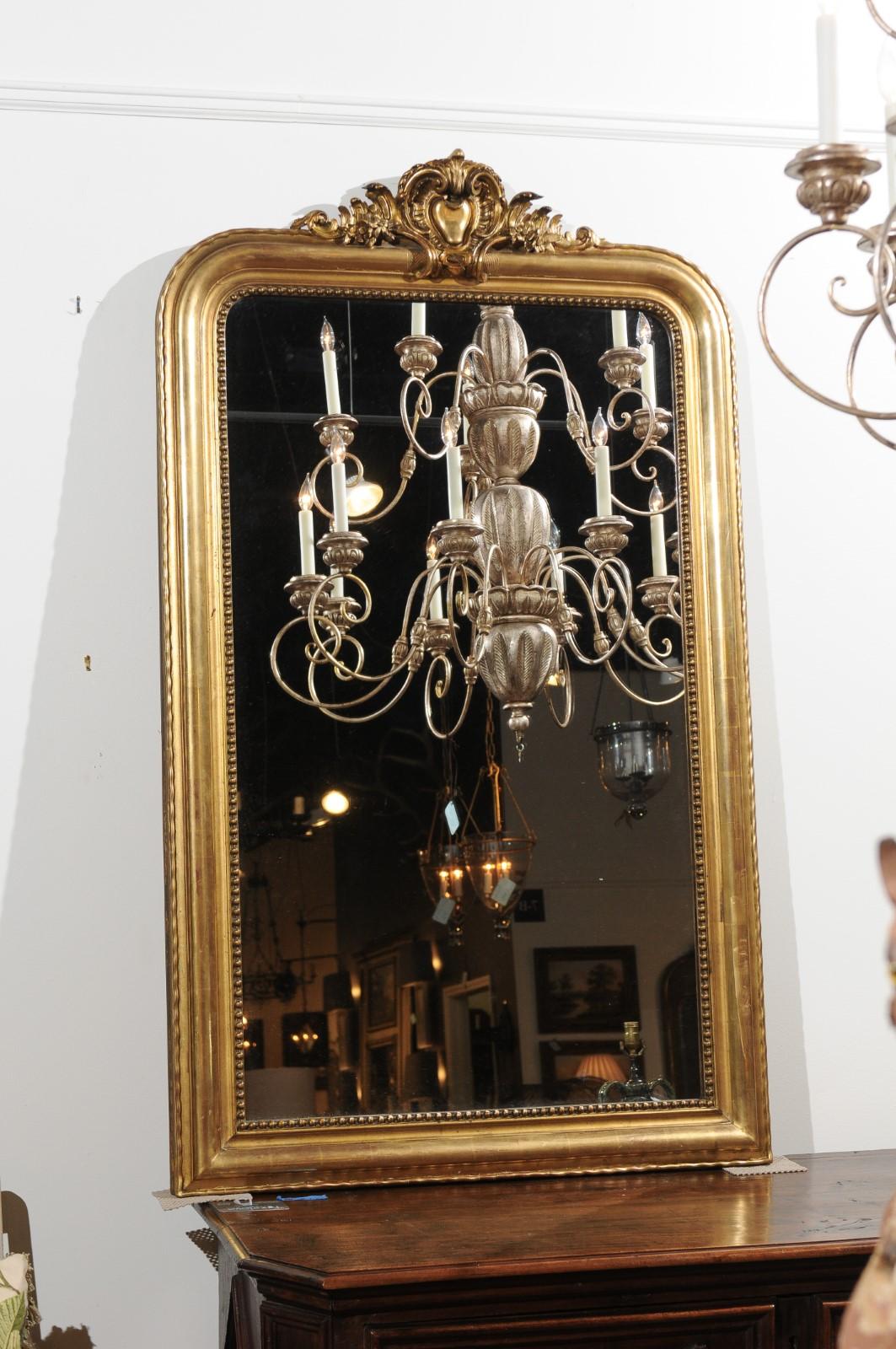 A French tall giltwood Louis-Philippe style mirror from the 19th century with hand-carved crest, beaded motifs and wavy pattern. This French Louis-Philippe style gilded mirror captures all of our attention with its exquisite hand-carved crest,