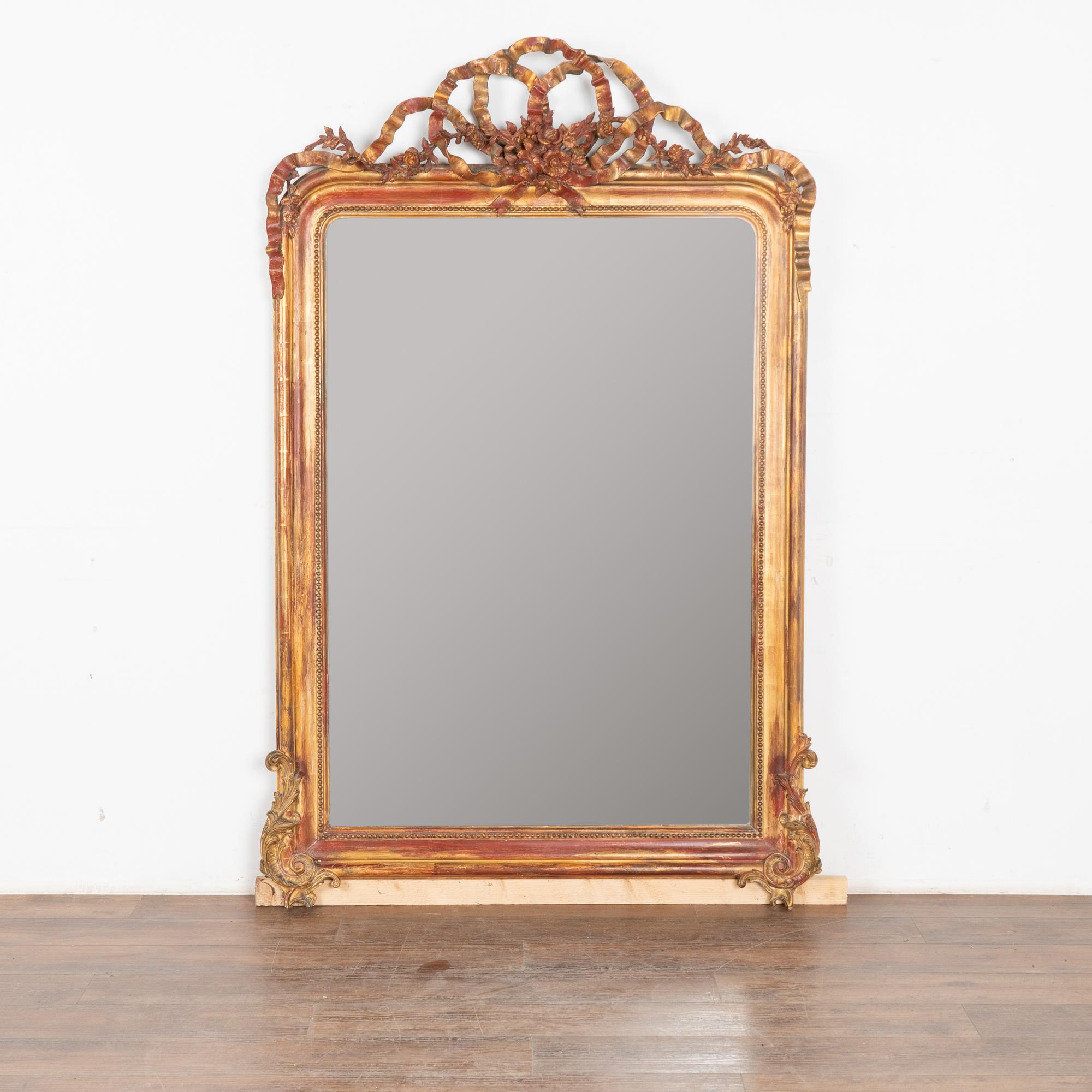 Questo straordinario specchio da 6' dorato presenta decorazioni intagliate in stile rococò, tra cui elaborati nastri con fiori. L'oro e il rosso brunito accentuano il bellissimo intaglio.
Venduto così com'è, in condizioni di utilizzo. È solido e