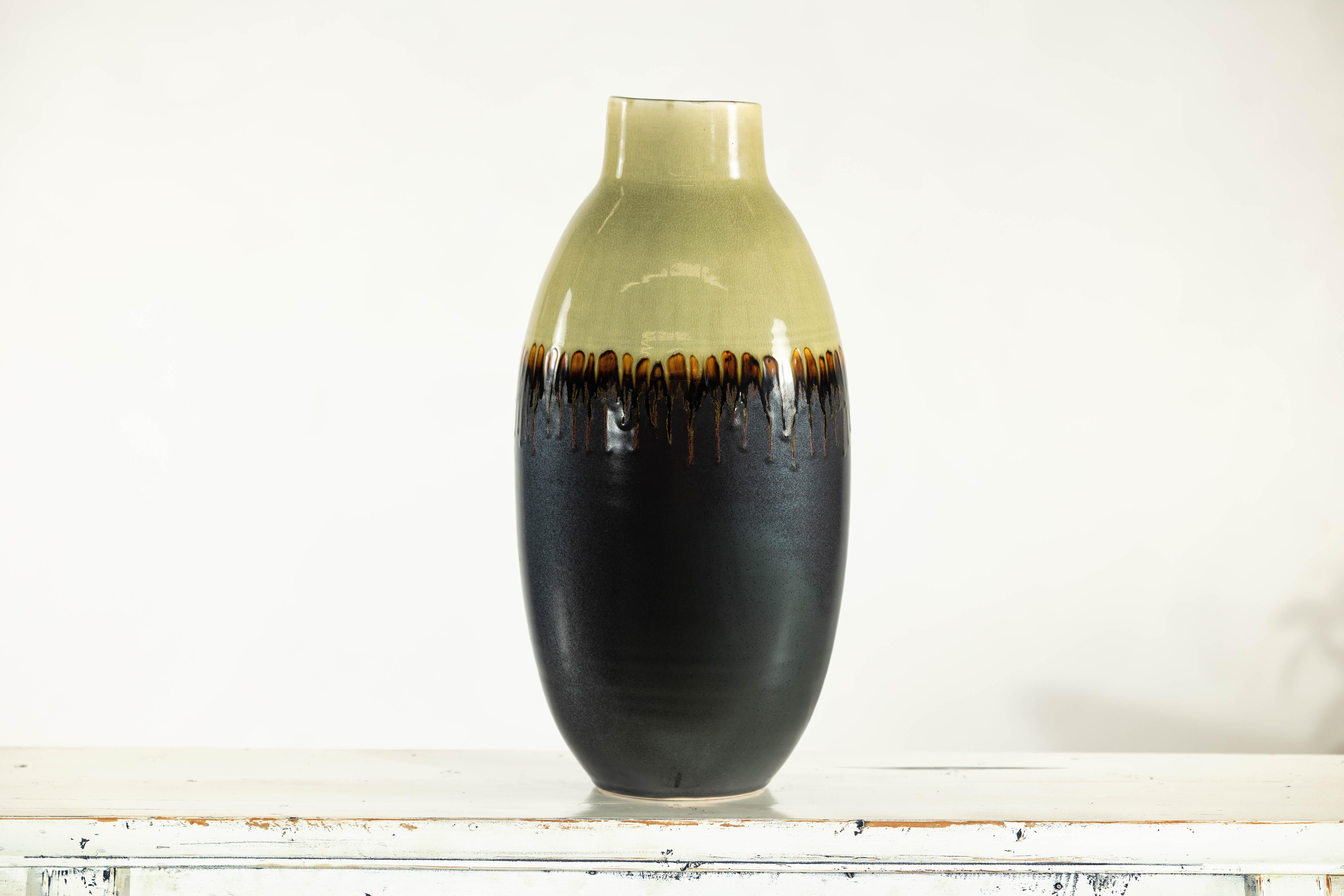 Questo alto vaso in ceramica vintage è caratterizzato da un sorprendente smalto bicolore con un caratteristico motivo a goccia. La parte superiore è smaltata in un tenue verde oliva, mentre il corpo passa a un marrone profondo, quasi nero. La