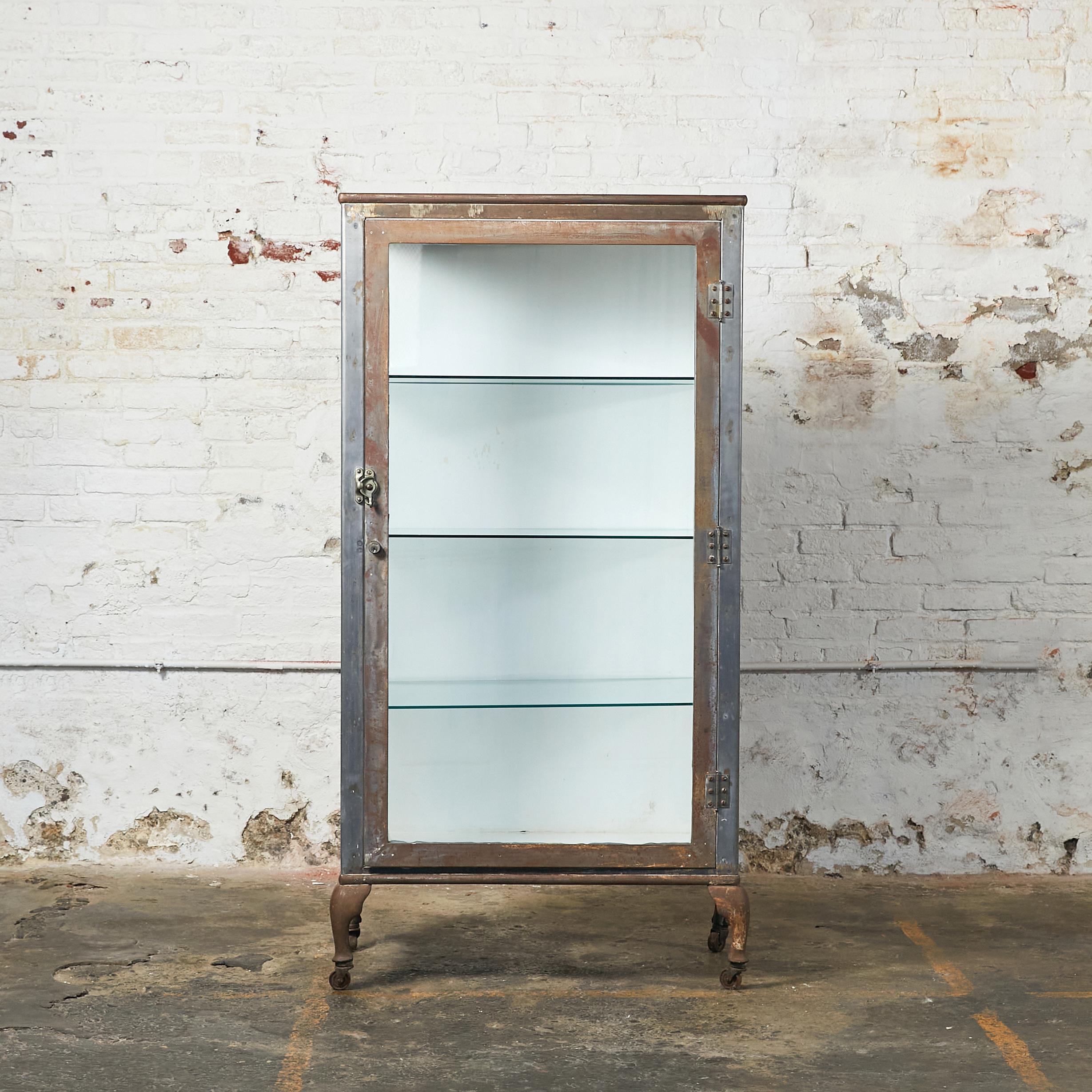 Tall industrial medicine cabinet. Metal frame door with clear glass and sides with ribbed glass inserts. The interior back and base are painted in white, and the shelves are made of glass. The cabinet rests on four curved shape metal legs with