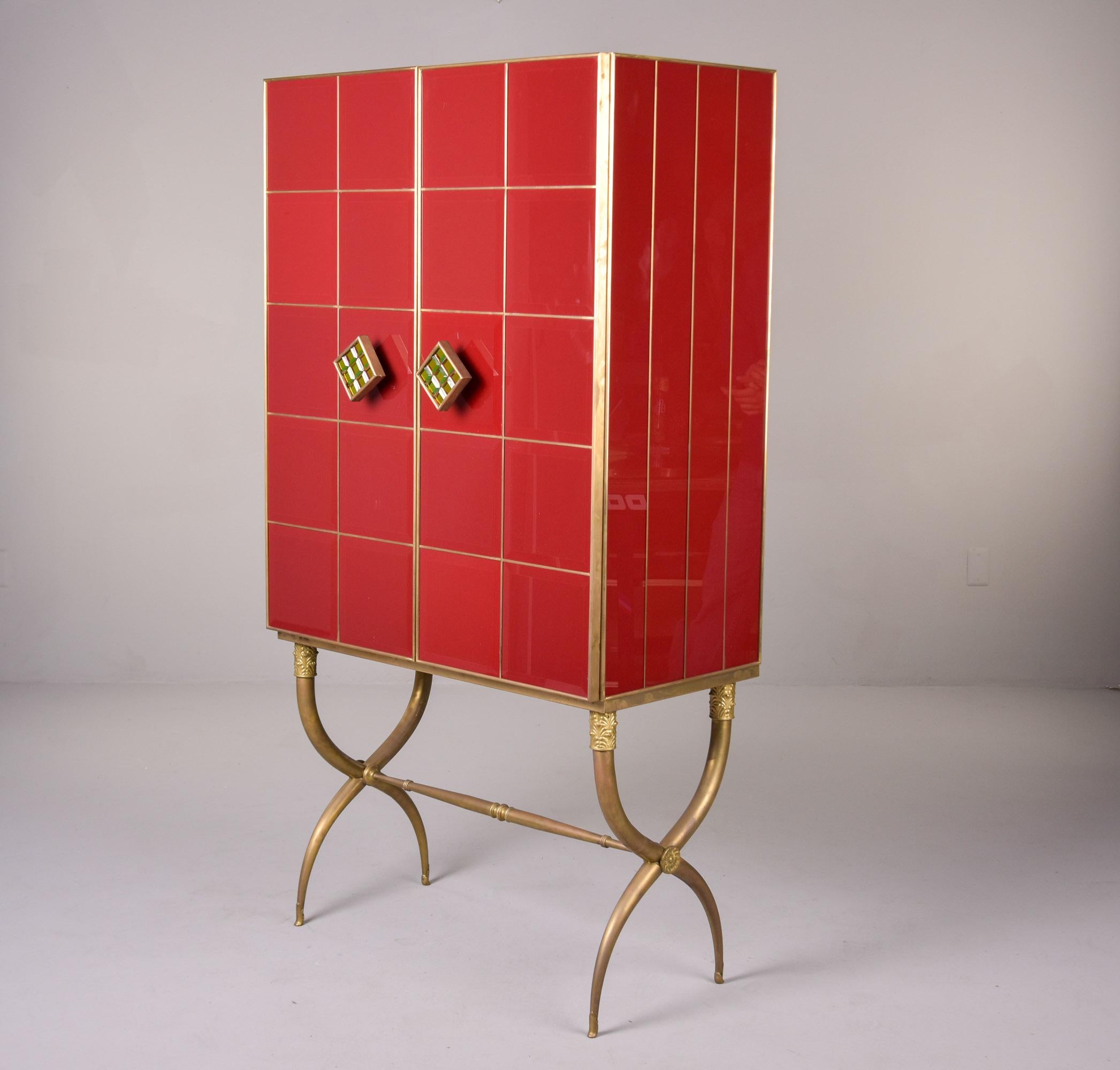 Found in Italy, this tall glass and brass-clad bar cabinet dates from the 1980s. Brass curule base has decorative details and supports a two door, glass and brass-clad cabinet. Glass panels/tiles have beveled edges and brass trim forms a grid