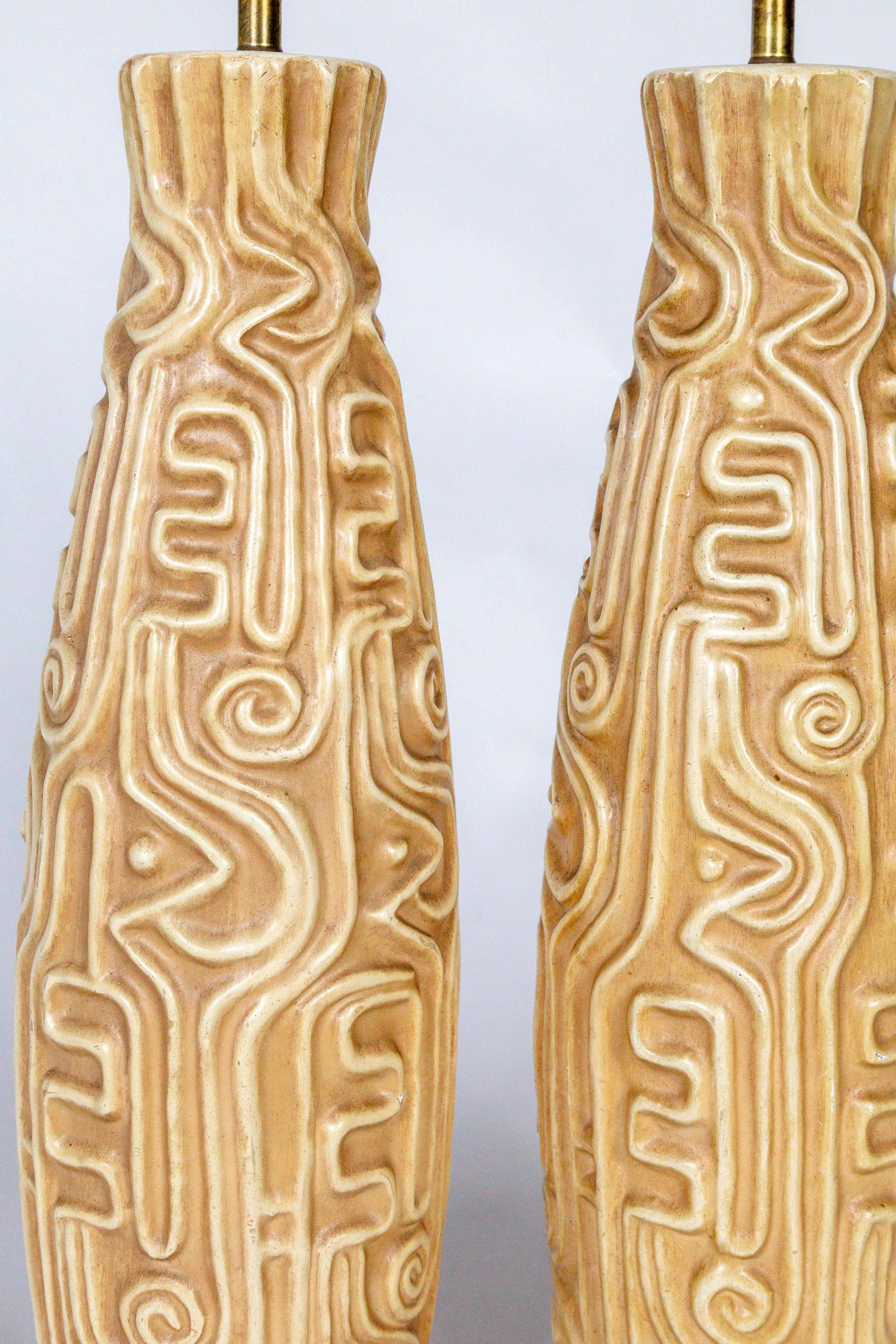 A pair of midcentury, American pottery lamps carved with high relief designs reminiscent of South American geoglyphs. Shaped tall and narrow and tapering slightly at the top in a light tan color, with a wooden base in a mahogany shade. With brass