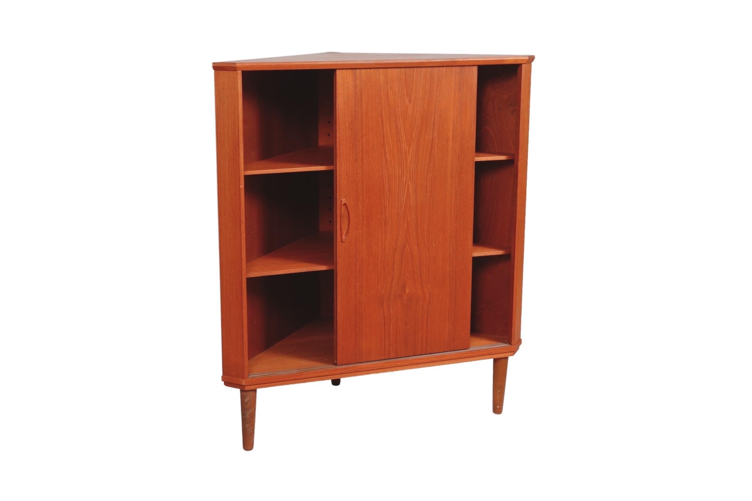 A tall mid century corner cabinet made of teak. Two adjustable shelves are housed behind two sliding cabinet doors with recessed handles. Stands on three round tapered legs.