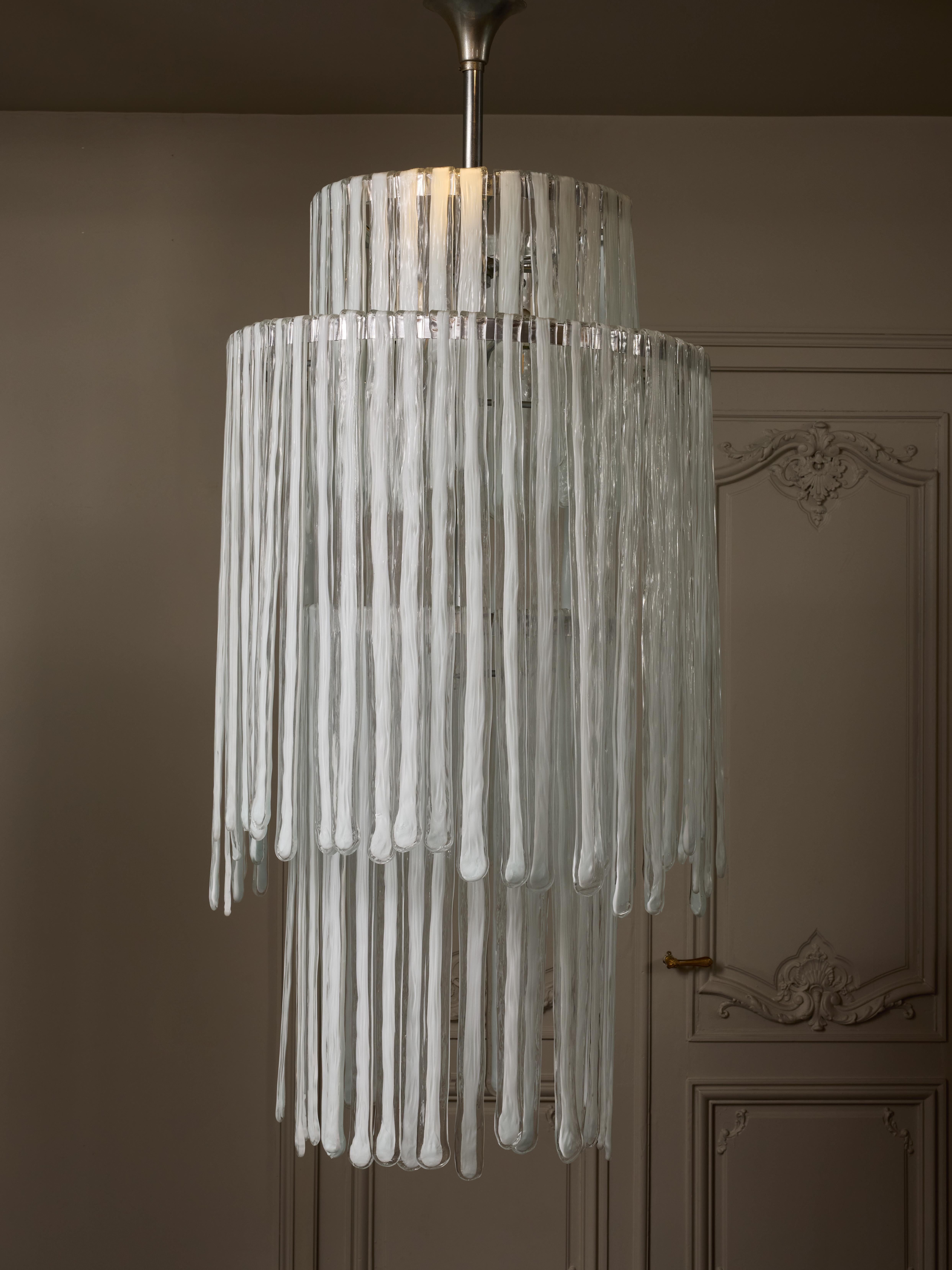 Alto lampadario vintage composto da una struttura in metallo da cui pendono decine di lastre di vetro di Murano trasparente fatte a mano con il bianco al centro, piegate in alto in modo da agganciarsi direttamente alla struttura.