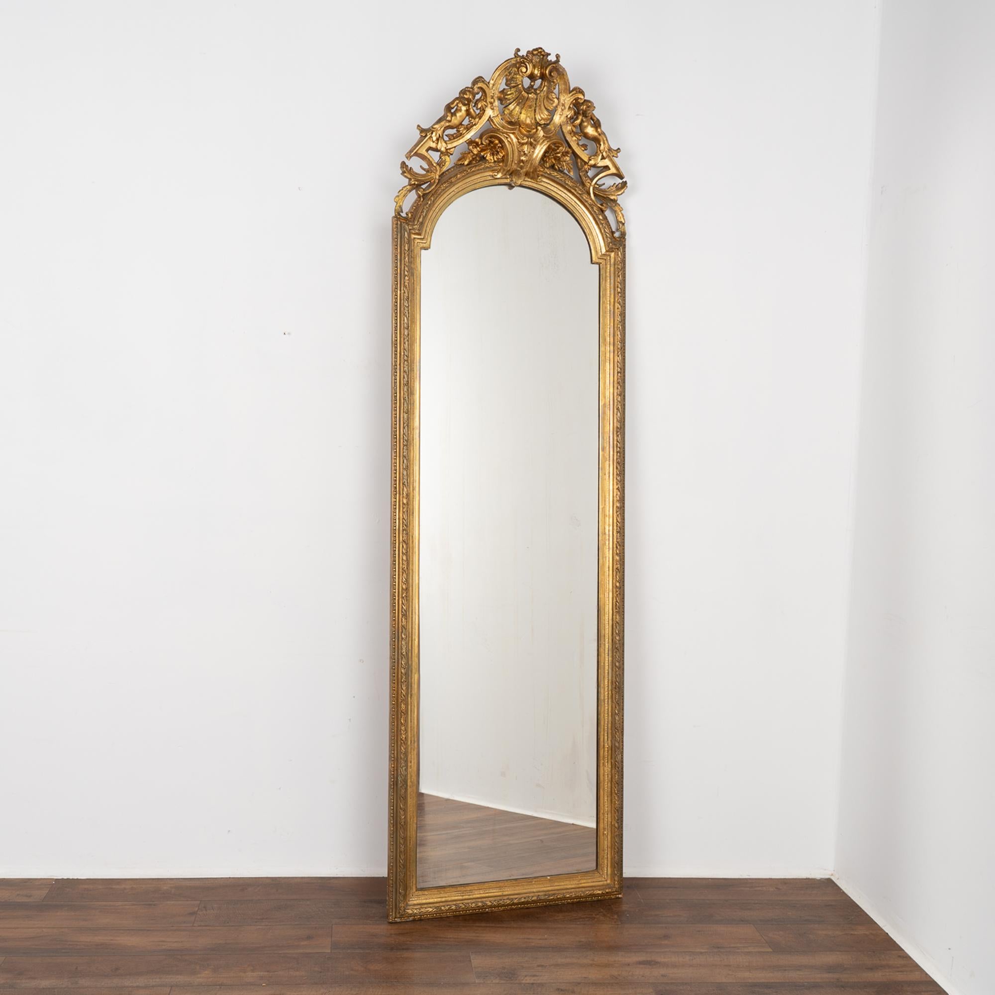 This stunning gold gilt mirror is over 8' tall and is crowned with carved flowers, two putti, leaves and flourishes.
There is expected age-related wear as seen where the gilt has been worn off, faded, chipped or cracked; some old repairs.
This tall