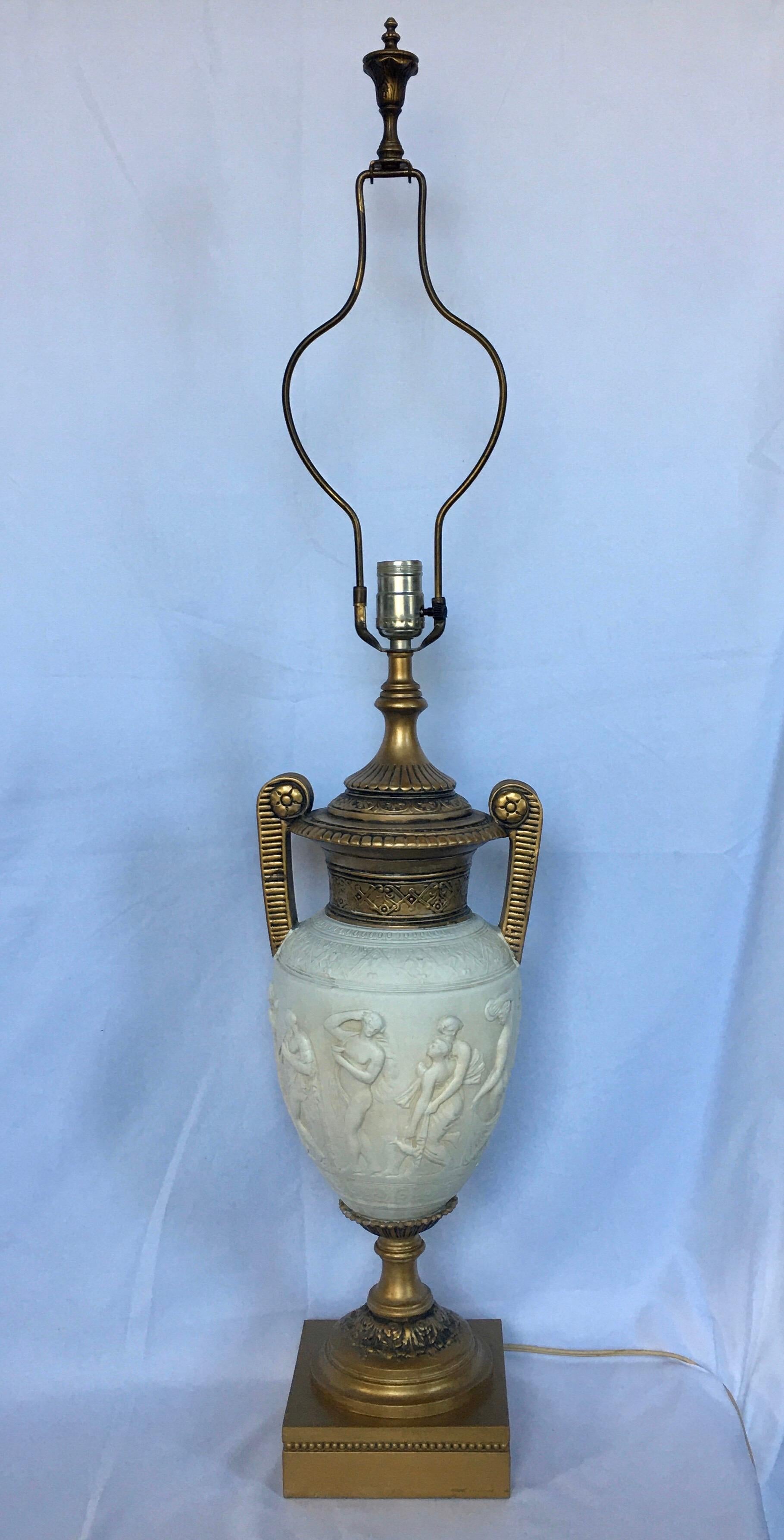 Tall neoclassical style urn table lamp with figurative relief. Matte cream ceramic urn features dimensional figures with gold painted metal handles and plinth base. Lamp shade not included.

Measures: Base width 6.25 inches.
Height to finial