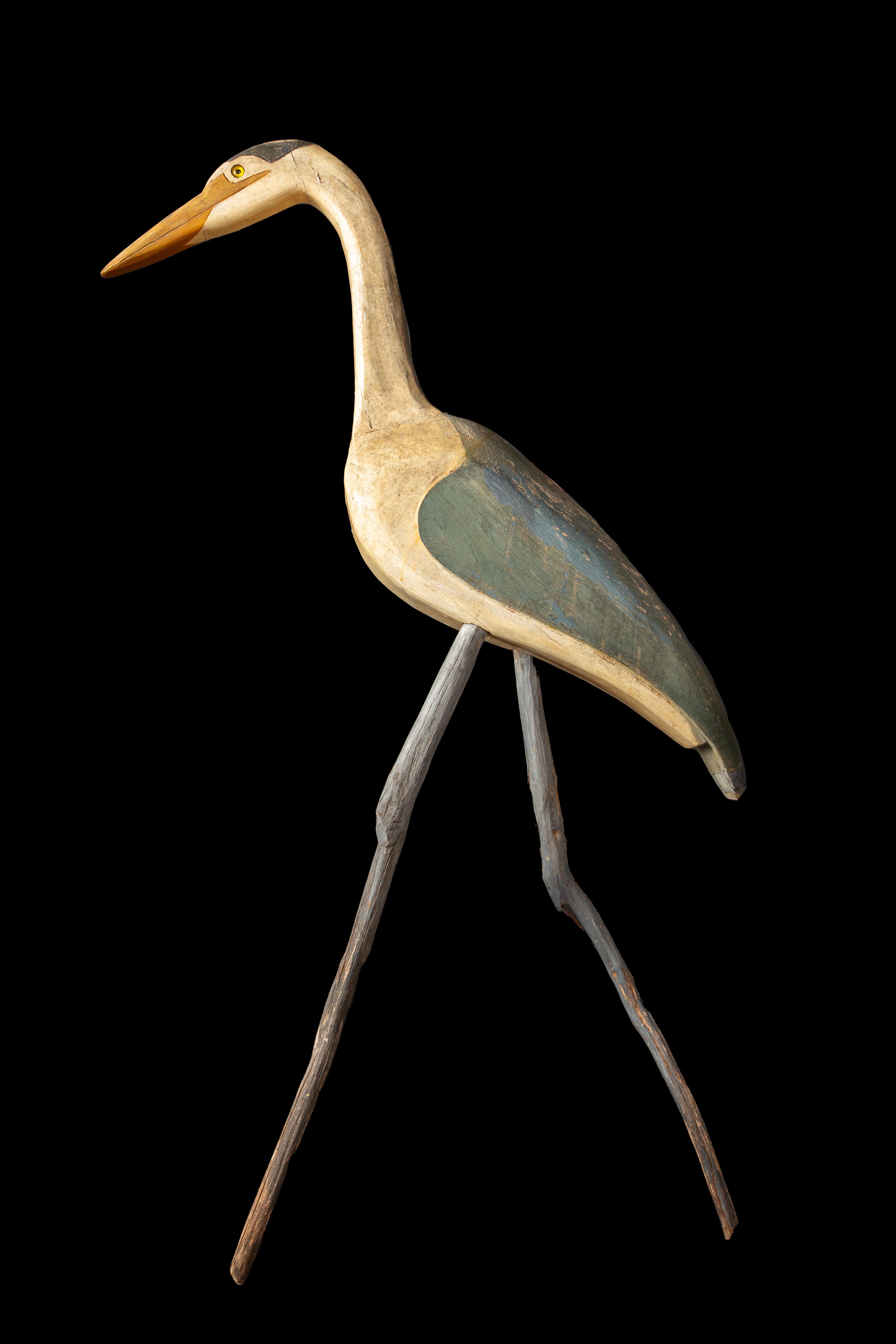 Tall Painted Carved Wood Folk Art Crane with Drift Wood Legs is an exquisite and captivating piece of artistic craftsmanship. Standing at an impressive height of 56 inches, this majestic crane emanates a sense of grace and elegance. The carvings on
