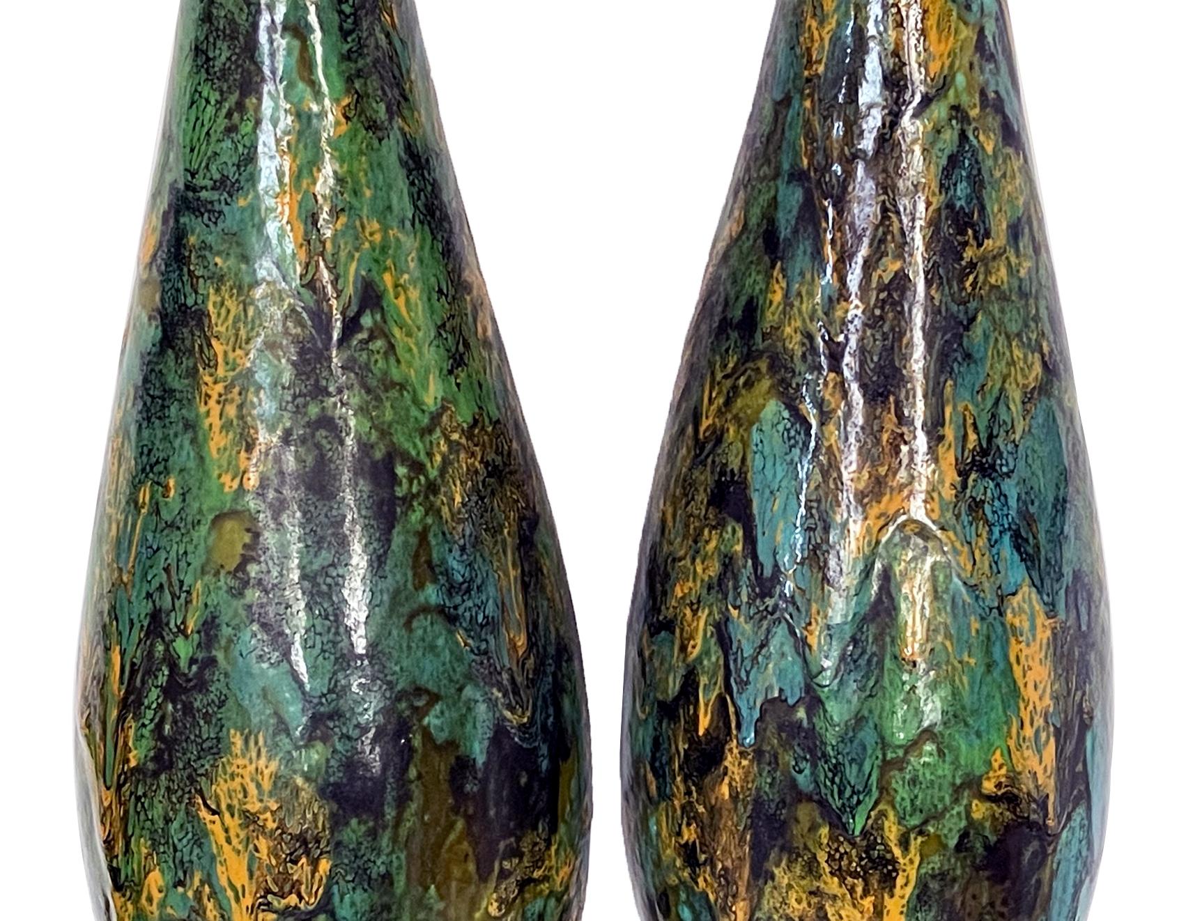 each tall slender lamp of bottle-form with black, yellow and teal drip glaze; raised on an ebonized metal base