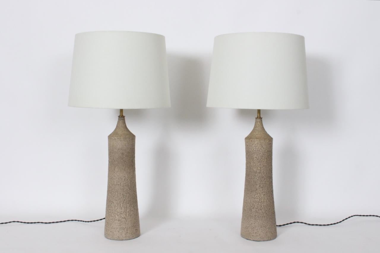 Pair Design Technics attributed textured earth tone Pottery Table Lamps.
Featuring handcrafted, smoothly coarse, slight hourglass matte glazed ceramic bottle forms with tapered tip in neutral Fawn coloration. 22.75H to socket. 18H to top of Ceramic.