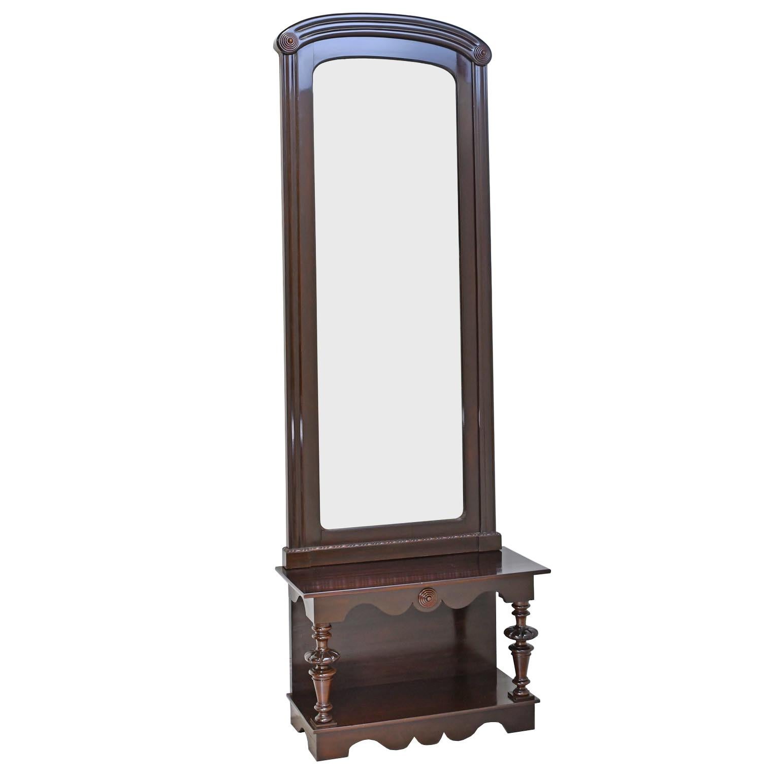 A tall and narrow hall or pier mirror in very dark polished mahogany with reeded molding and arched top ending in bullseye rosettes resting on a low console supported by columns composed of bell-and-baluster turnings, whose apron and plinth are