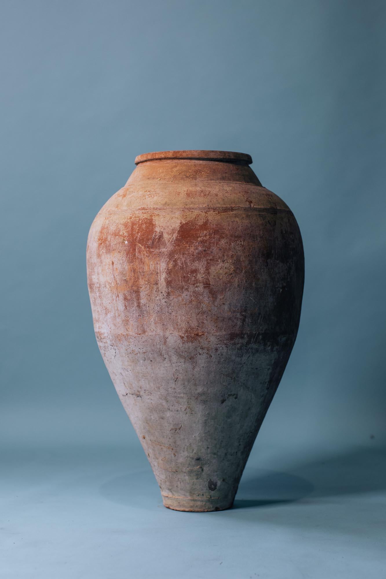 A striking early 20th-century Mediterranean storage jar, hand-thrown from red terracotta clay and fired to reveal its natural tonal shifts of rose, umber, and smoke-grey. Its tall, narrow form and softly tapered base suggest it was once used to