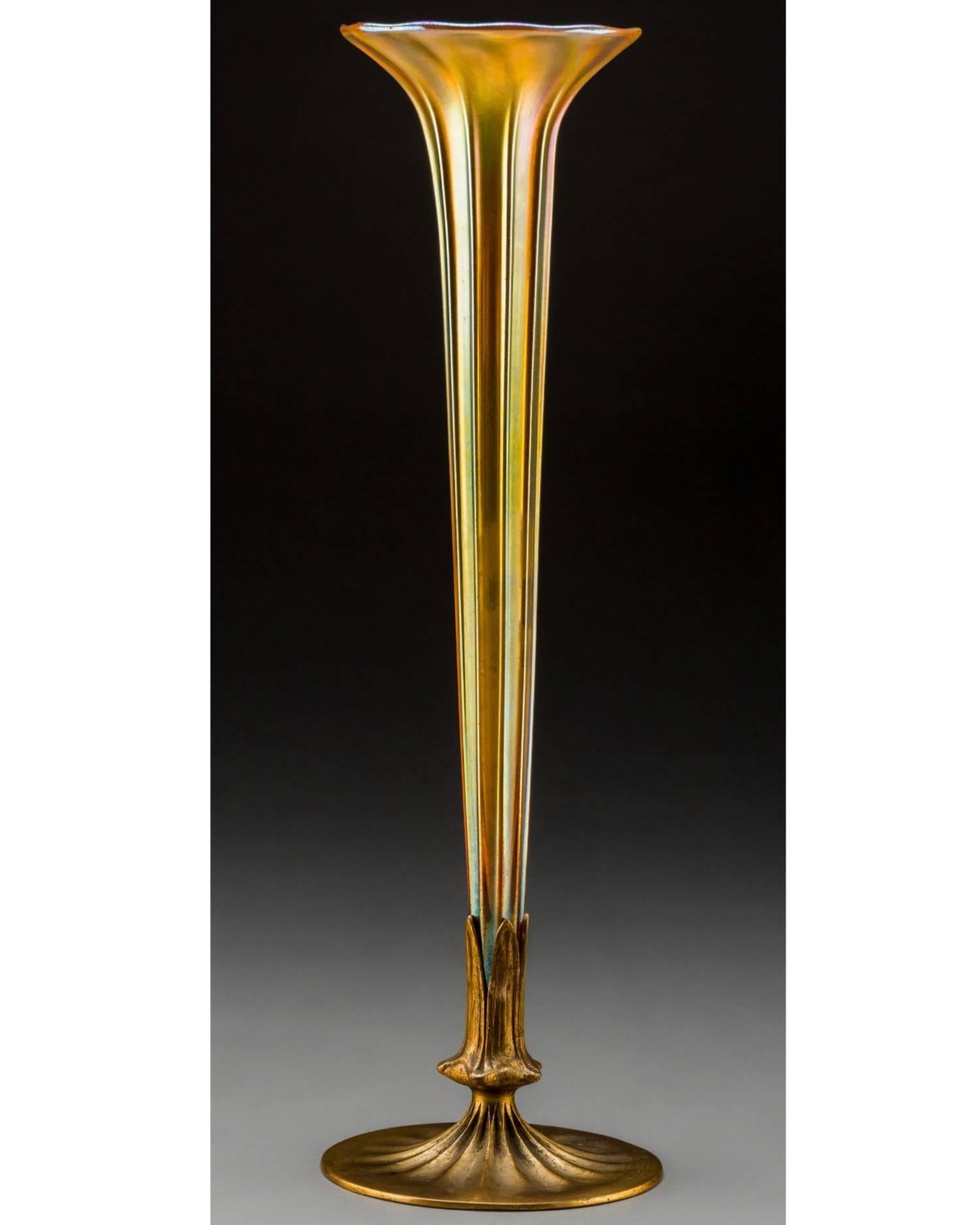 A Tiffany furnaces gold favrile glass and gilt bronze trumpet vase. Favrile glass is ribbed with iridescence, circa 1920-1928. 

Base stamped LOUIS C. TIFFANY FURNACES, INC., 158, with logotype; Glass engraved L.C.T., Favrile. 

Condition: Excellent