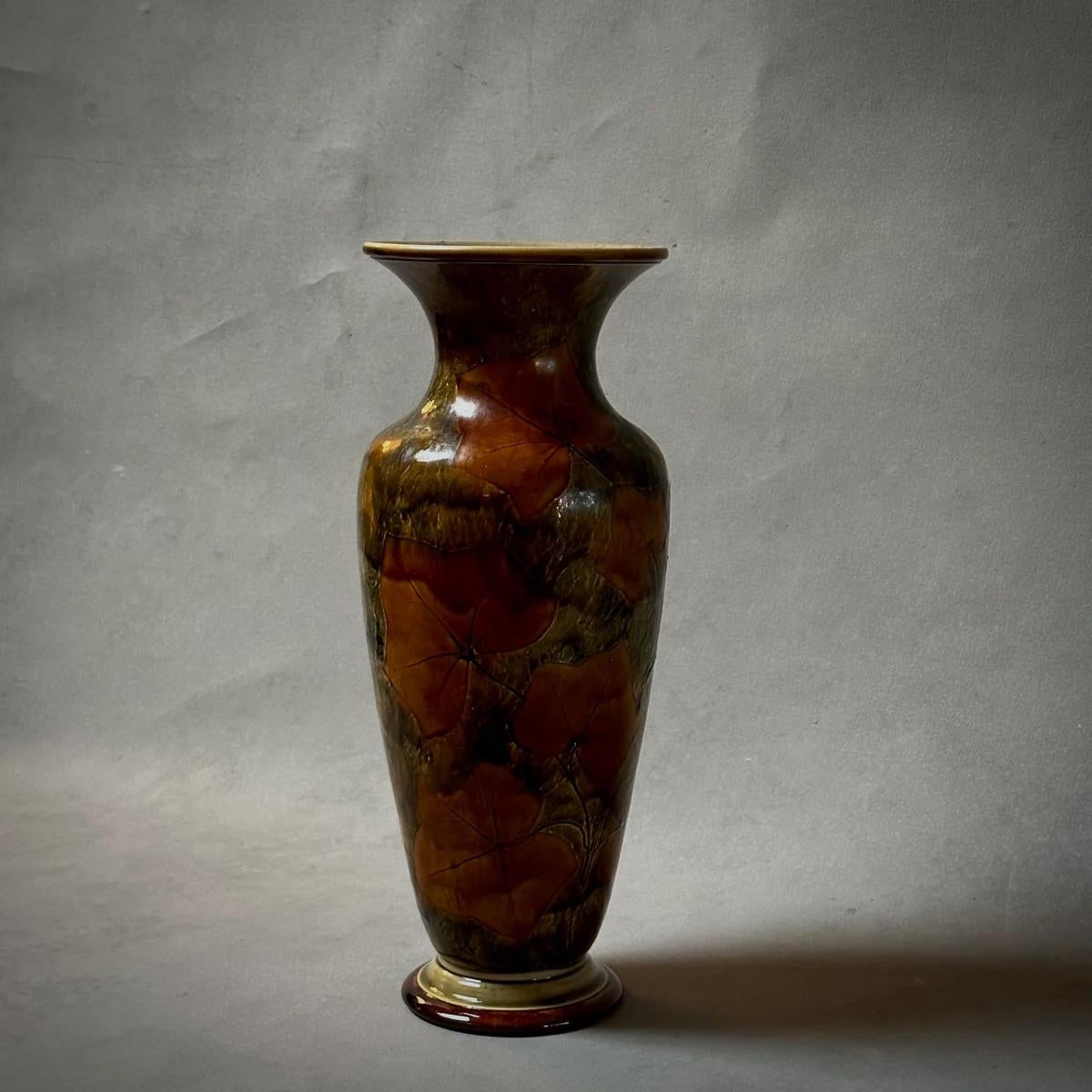 Este jarrón de Royal Doulton, de principios del siglo XX, muestra un sofisticado motivo de hojas en una paleta de tonos caqui, oliva y musgo. La abertura acanalada complementa con gracia su base circular elevada, formando una silueta equilibrada que