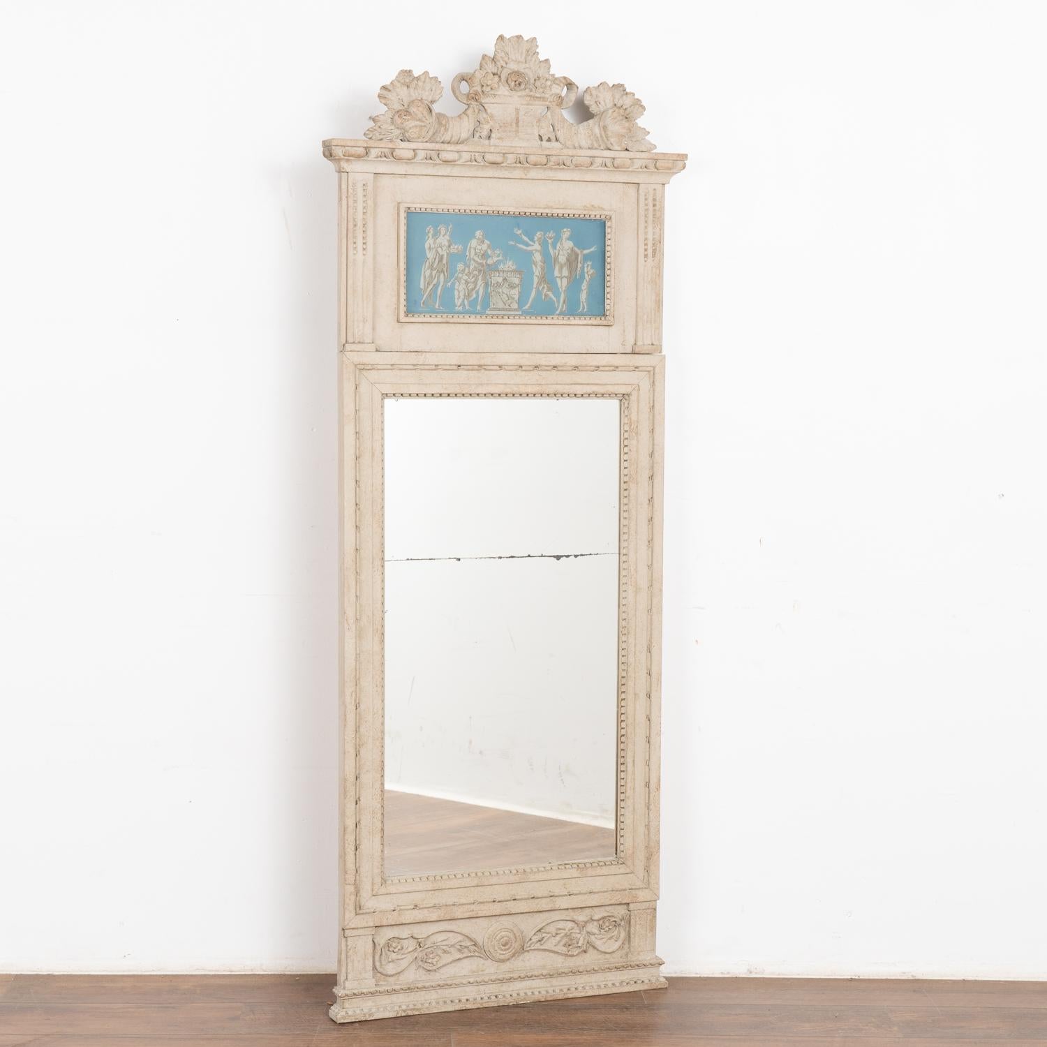 Tall decorative trumeau mirror with carved accents. Upper section portrays Greek figures against a blue painted background.
The antique white painted finish is lightly distressed creating a refined grace fitting the age of the mirror.
Mirror glass