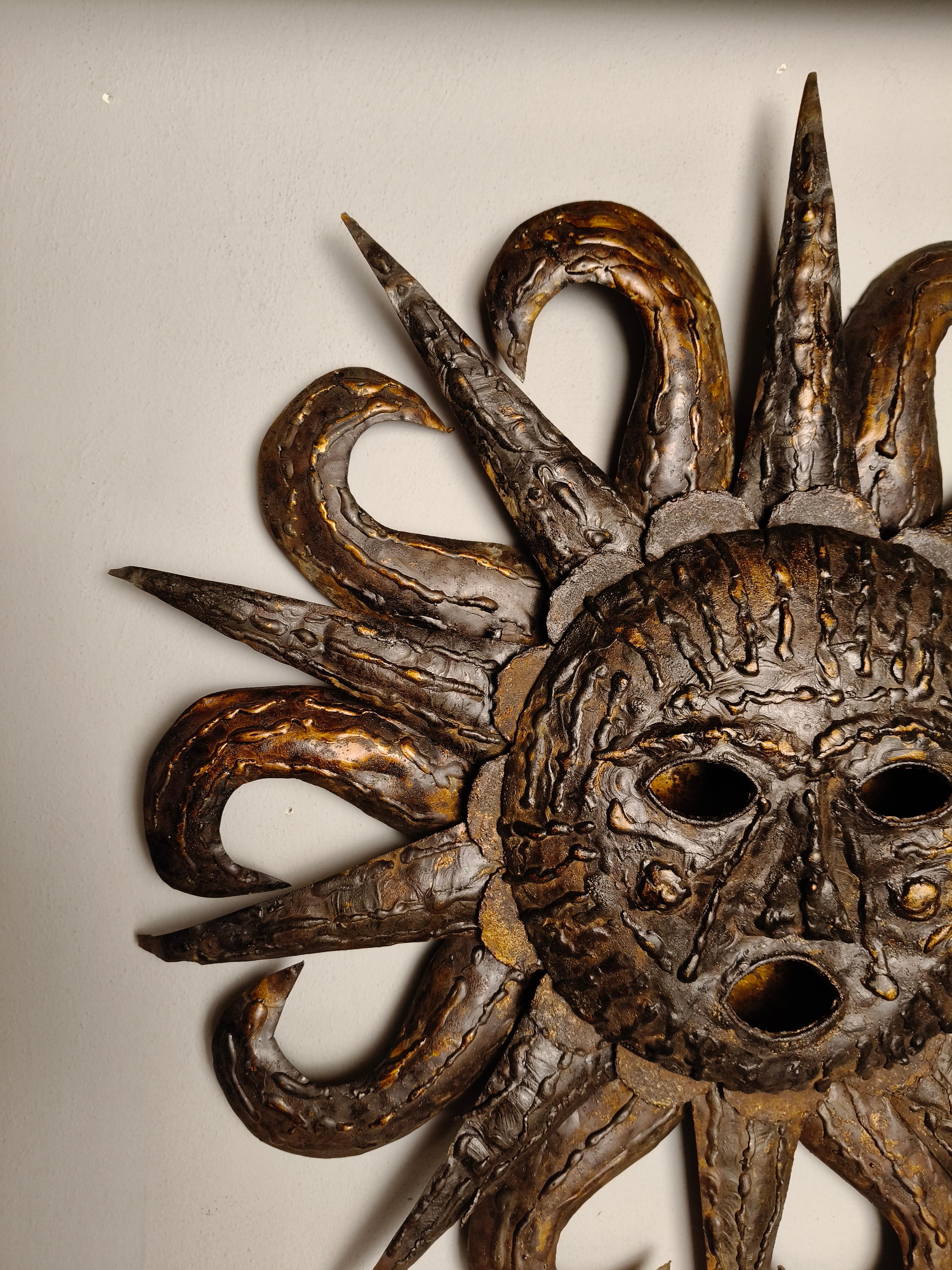 Escultura mural de acero martillado de mediados de siglo de Talleres Emaús, en Cuernavaca, Morelos, México. El rostro del sol tiene los ojos hundidos y la boca abierta. Tiene dos mejillas redondas. Diez rayos rectos y diez rayos curvos, hasta un