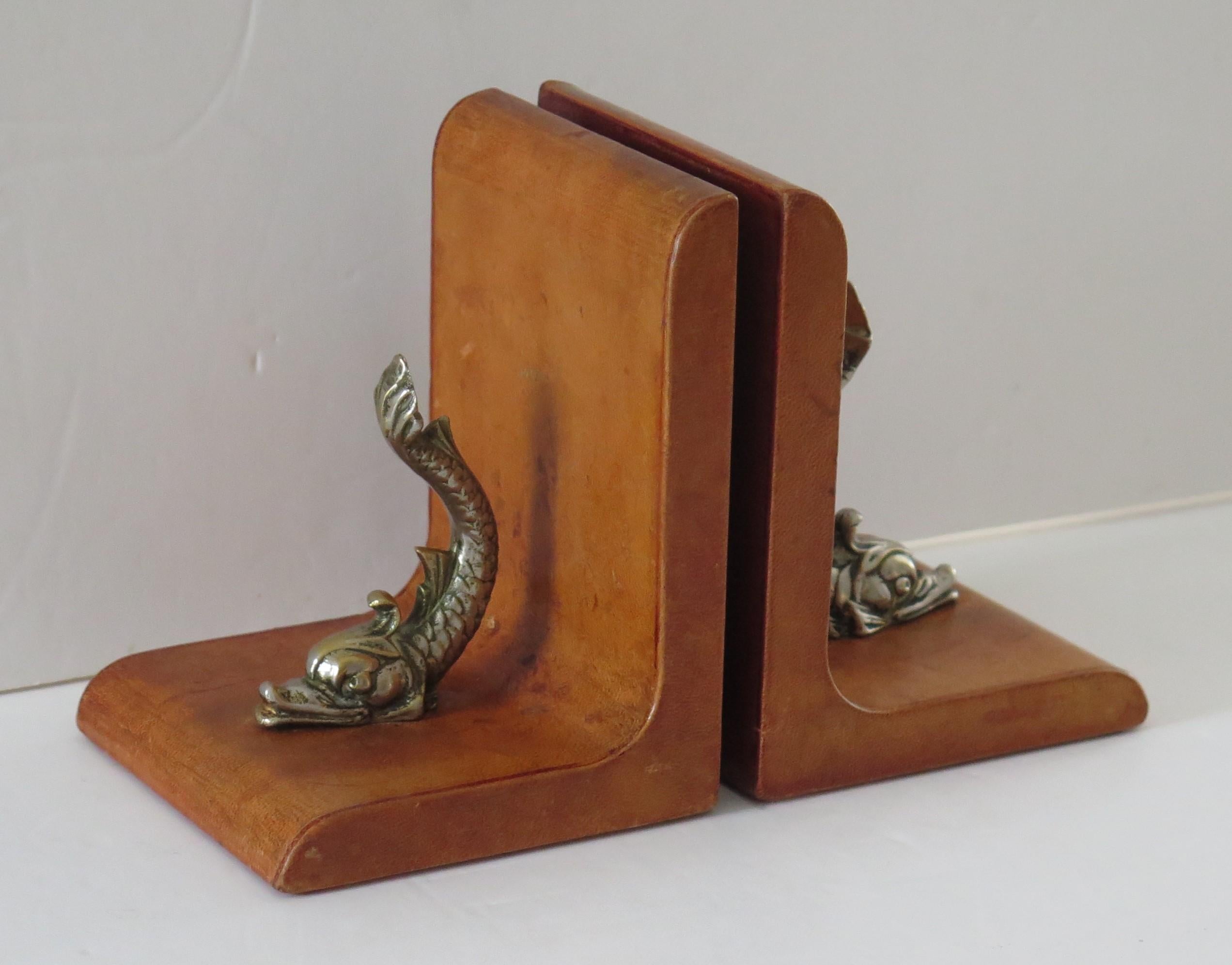 These are a beautiful, pair of Tan Leather book ends, mounted with gilt bronzed metal dolphins, which we date to the mid 20th century.

The bookends have been beautifully modelled in a tan coloured leather with each piece having a mounted stylised