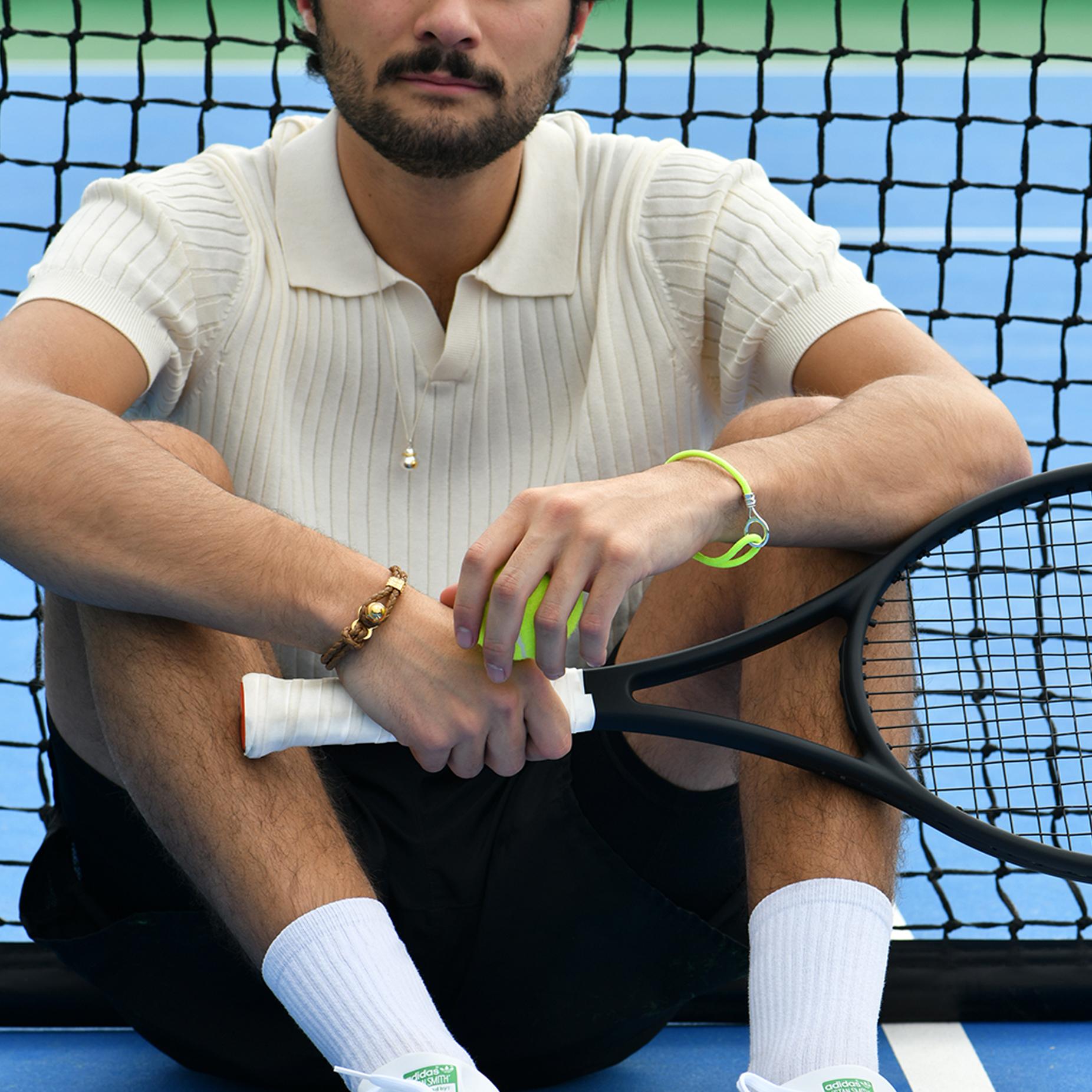 The TANE Tennis Ball Gold Bracelet from the #TANETENNIS collection is handcrafted in leather with an 18K Yellow Gold button closure system. At the center of the piece is sculpted in silver a tennis ball, accompanied by a clamp with engraved net