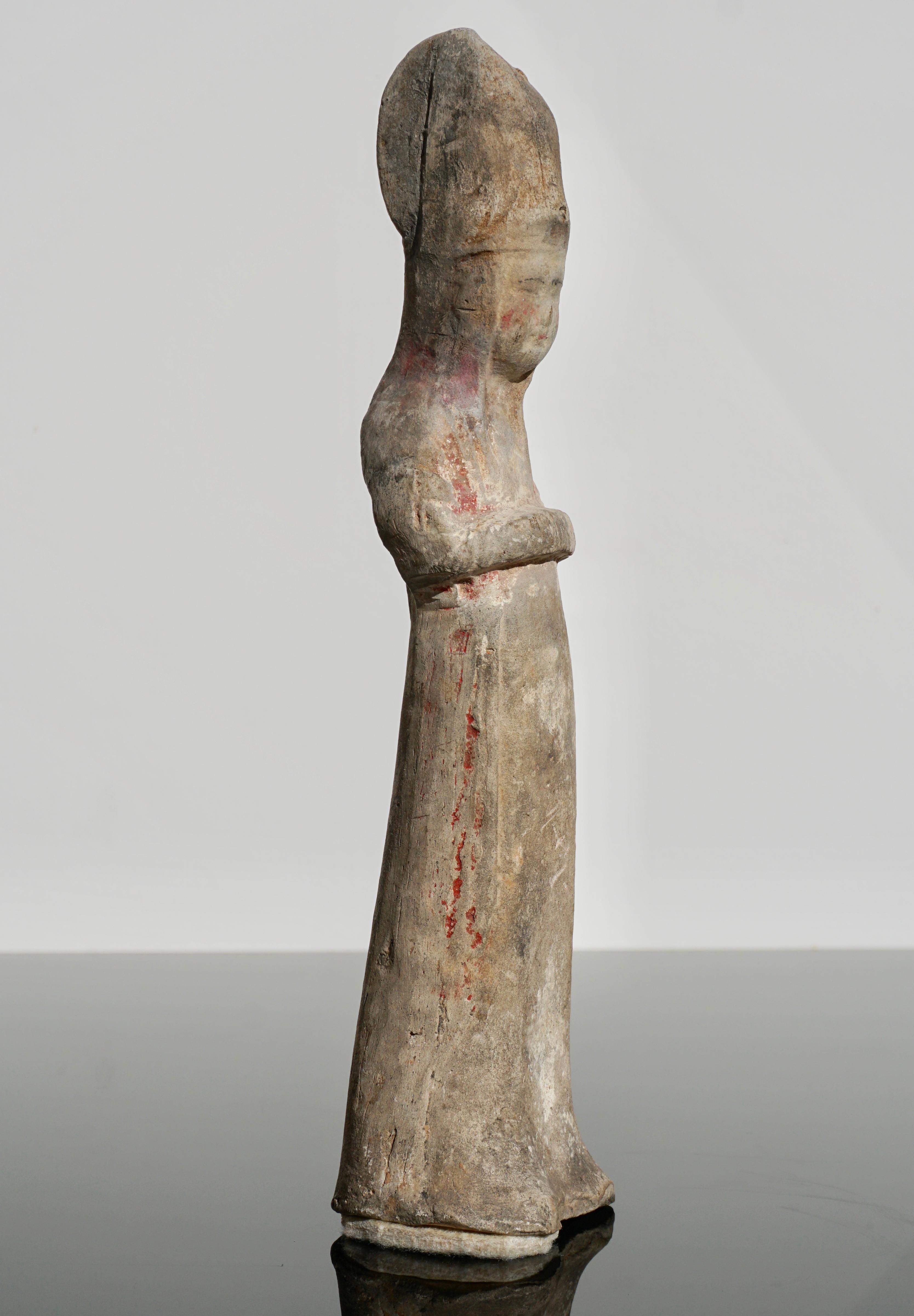East Asia, China, Tang Dynasty, circa 618 to 907 CE. 

A beautiful mold-formed pottery tomb attendant in the form of a female noblewoman. She wears long robes and stands with her hands folded in front of her. On her head is a conical headdress or