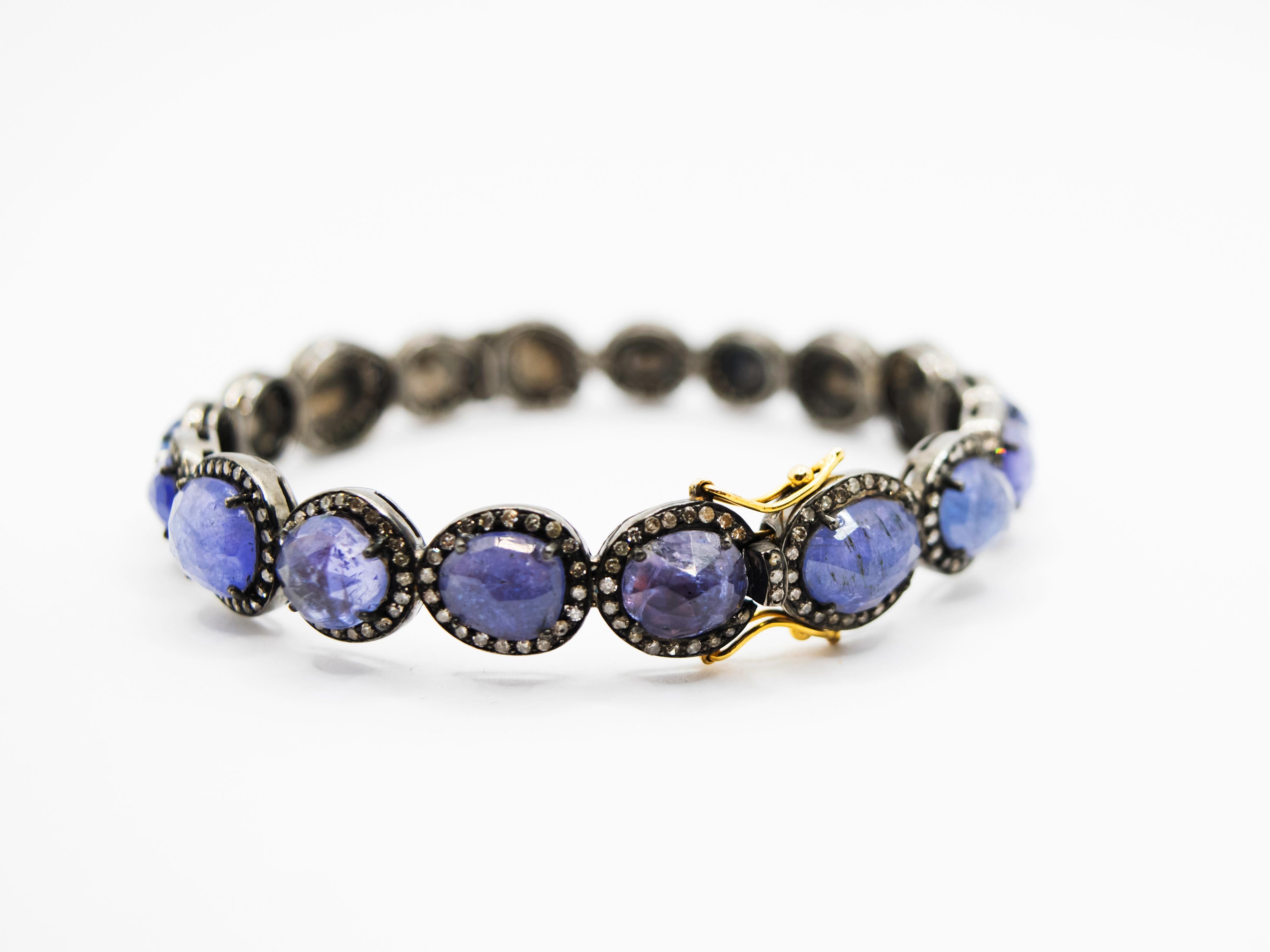 Bangle bracelet in grayed silver and 18 Kt yellow gold clasp.
Rigid, this bracelet is adorned with 18 irregular and different oval cut Tanzanite in very particular blu/ purple color.
Every stone is surrounded by gray diamonds. The diamonds have