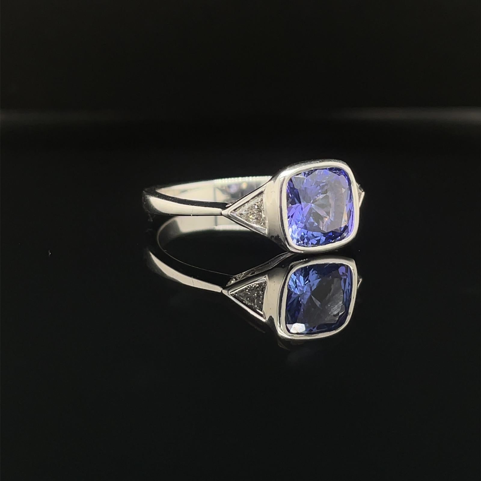 A tanzanite and diamond three stone engagement ring in 18 karat white gold.

This classic and contemporary ring is set to its centre with a distinctive lively blueish purple cushion cut tanzanite of 1.46 carats, mounted into a polished rubover
