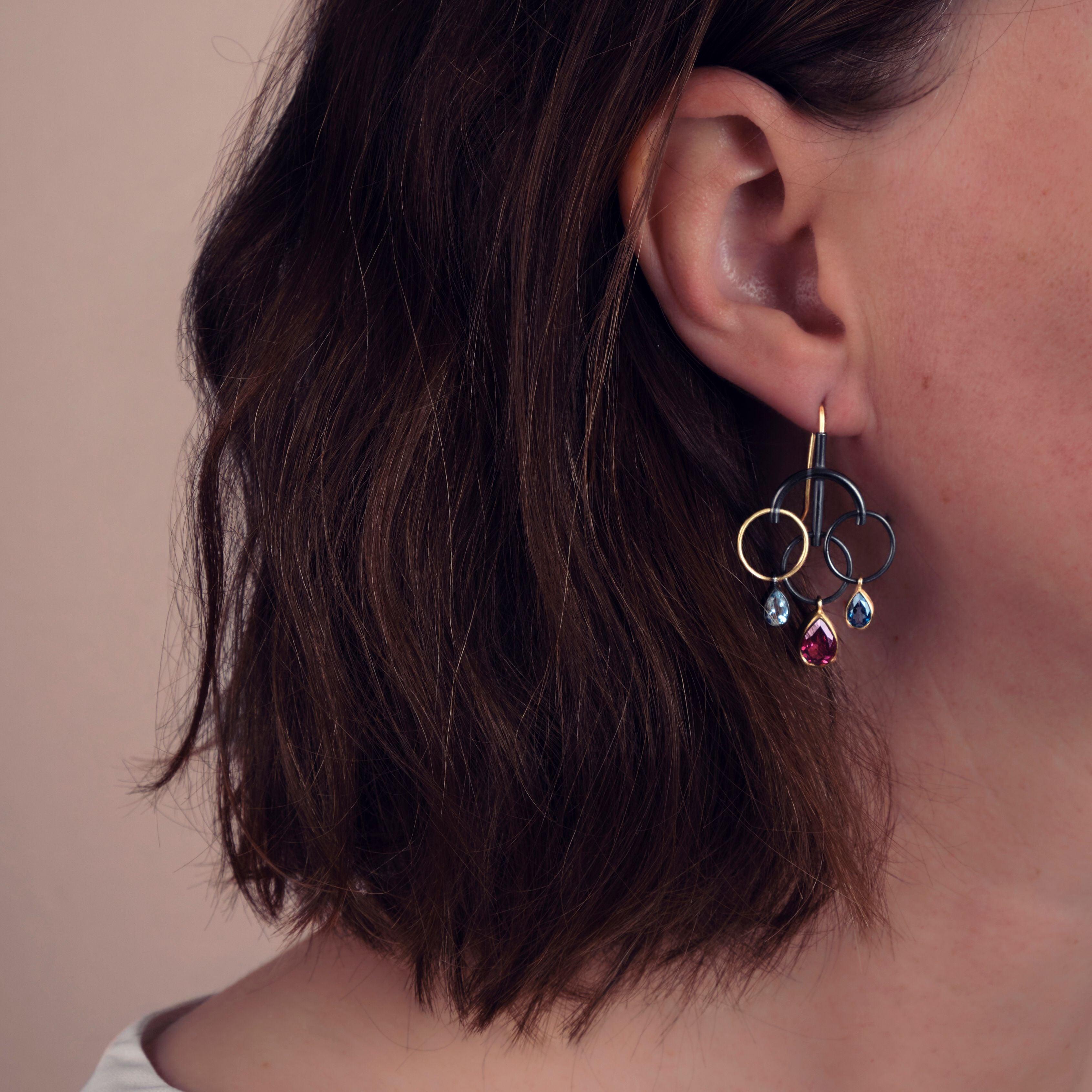 These very contemporary oxidised sterling silver and 18K gold drop pierced earrings playfully feature a brilliantly balanced combination of faceted tanzanite, aquamarine, London blue topaz, pink amethyst and rhodolite garnet. 
These earrings are