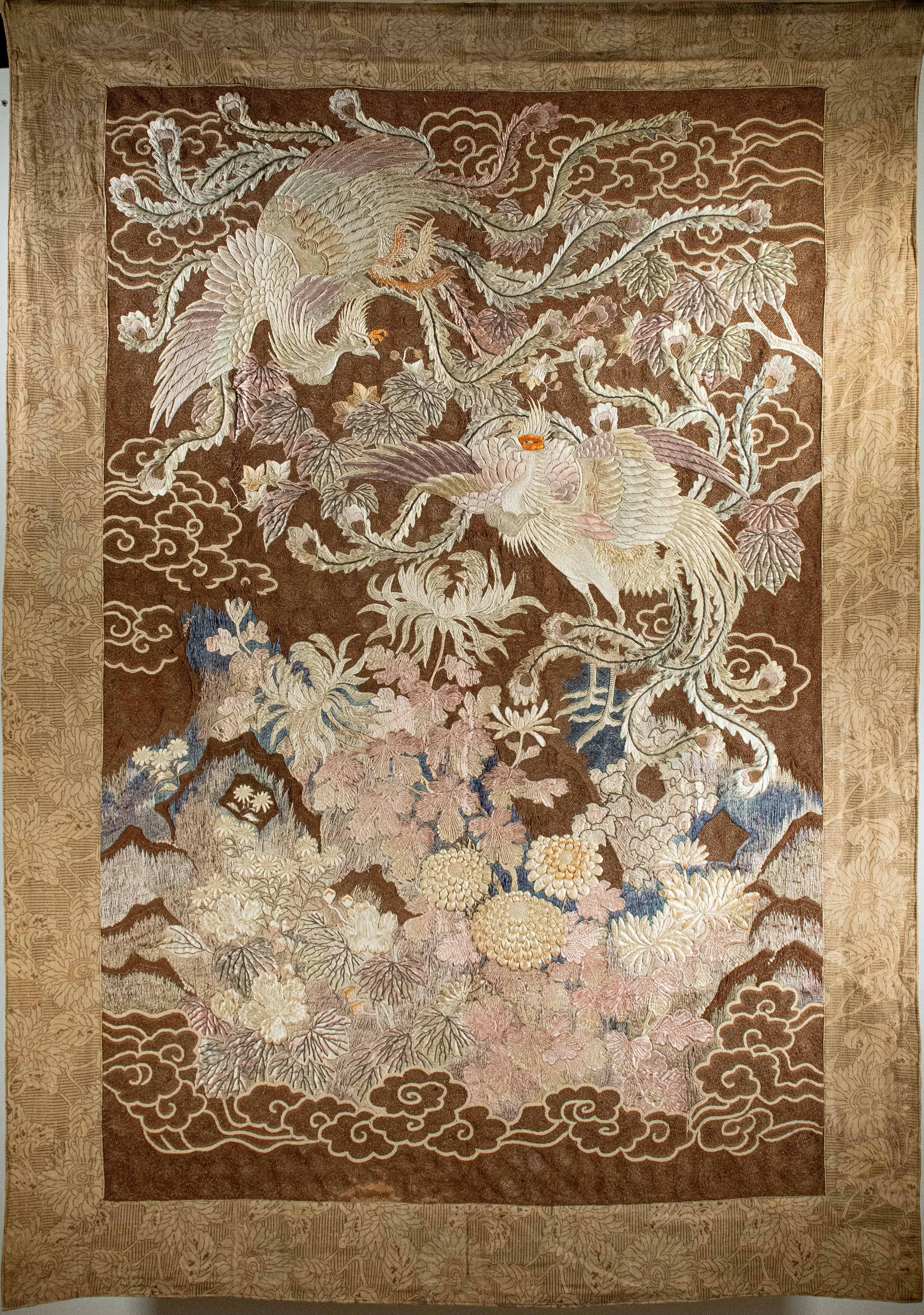Tapestry embroidered with polychrome silk threads, depicting two phoenixes flying among the clouds, above a garden of colored chrysanthemums. The frame and the back of the tapestry, protecting the embroidery, are decorated with a delicate floral