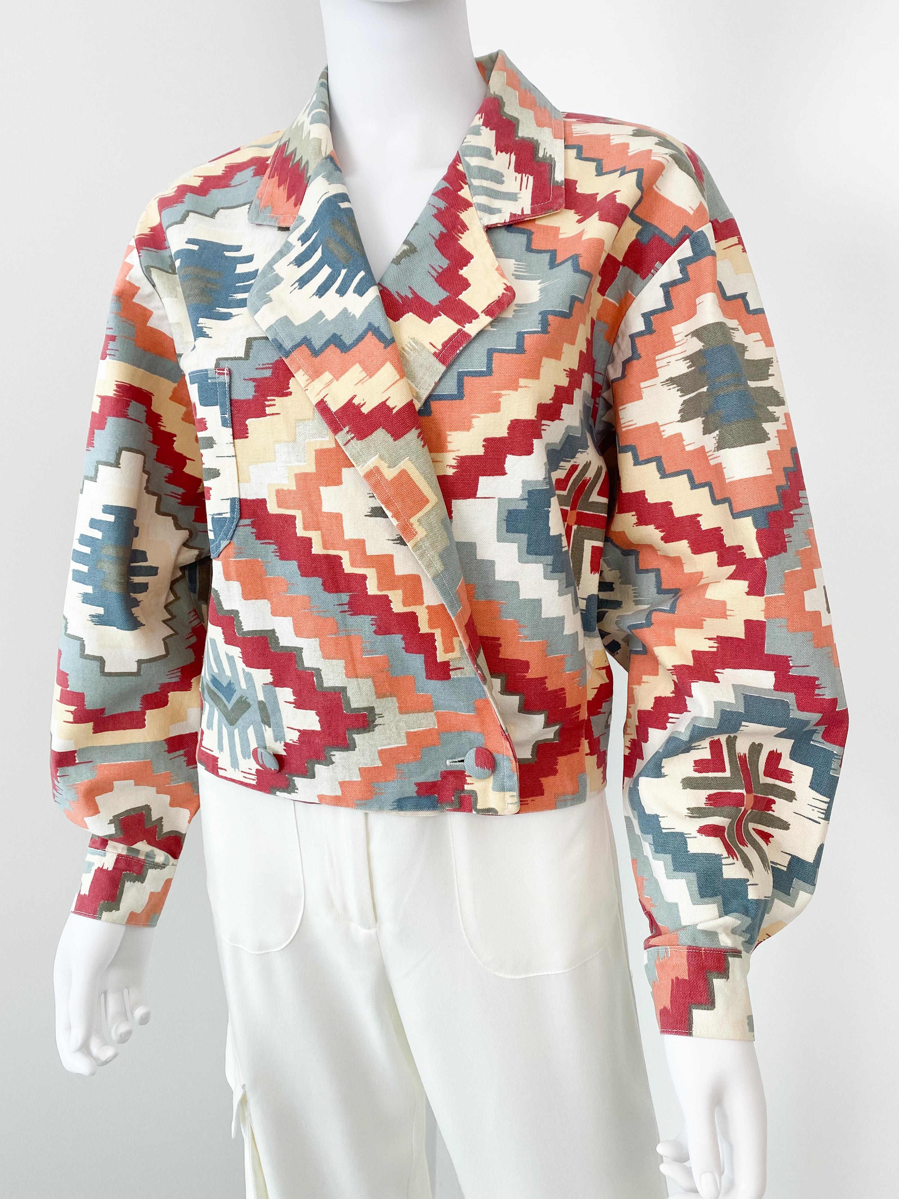 This vintage double-breasted bomber jacket from the 1980s features a multicolor ethnic-inspired print on tapestry-like cotton blend, long sleeves with cuffs, and covered buttons fastening on the front. The slightly stiff fabric highlights the