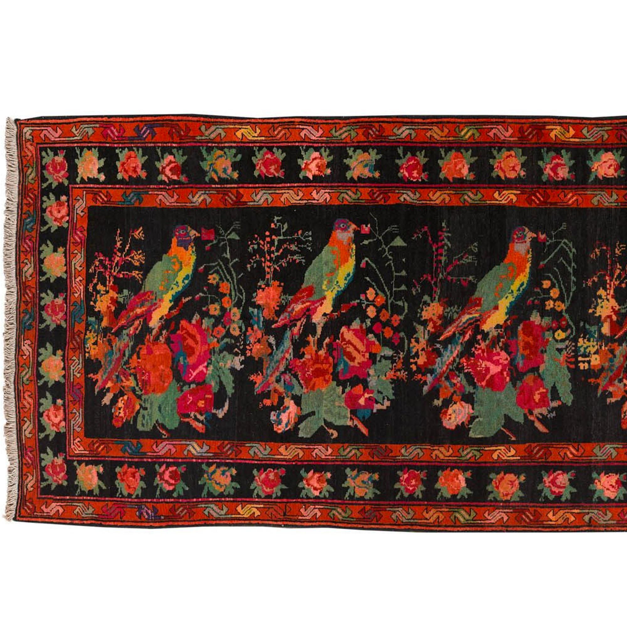 Der Teppich mit einer lebendigen, malerischen Ästhetik. Die Oberfläche zeigt florale und naturalistische Motive mit großen bunten Vögeln in der Mitte und einer reich verzierten Bordüre. Die Grundfarbe ist schwarz, mit leuchtenden Schattierungen von