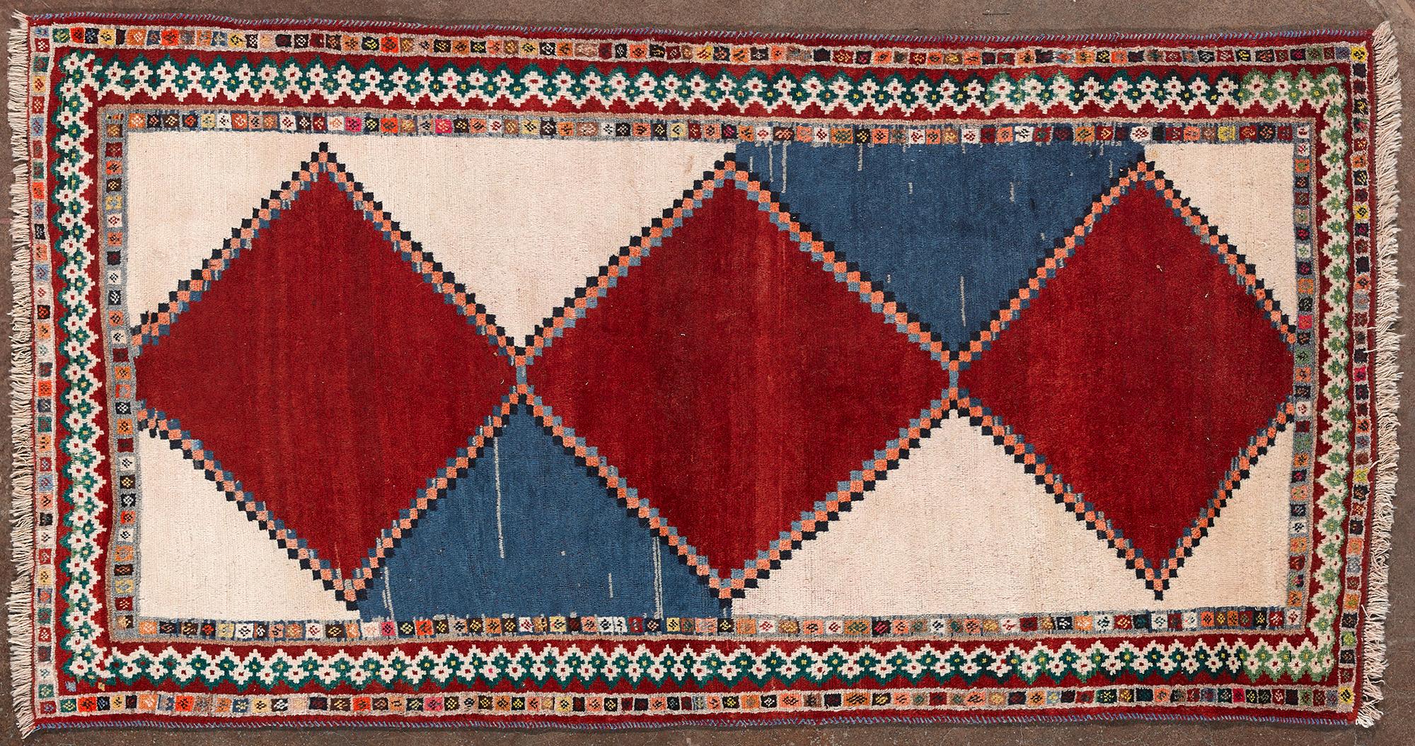 Alfombra tribal anudada a mano de origen caucásico o persa, fabricada a finales de los siglos XIX y XX. Presenta un diseño geométrico con tres grandes rombos rojos centrales alternados con secciones azules y beige. El borde decorado con motivos