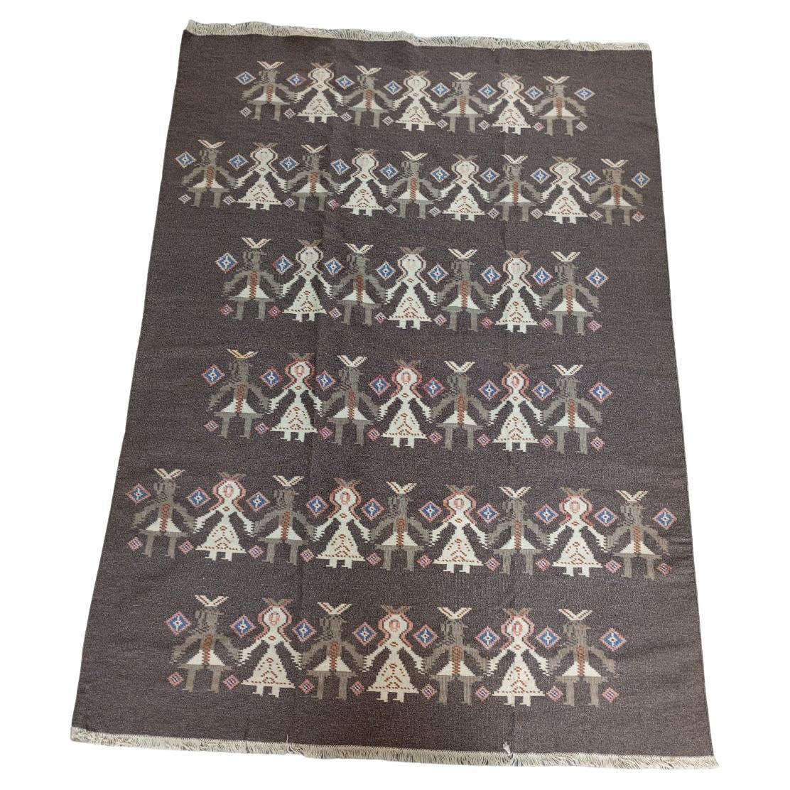 Tapis Kilim marron des années 1970 avec des figures stylisées disposées en rangées régulières  et horizontales, sur fond brun. Le fond du tapis est d'un brun chaud et uniforme, qui sert de base aux motifs. Les figures des garçons et des filles sont