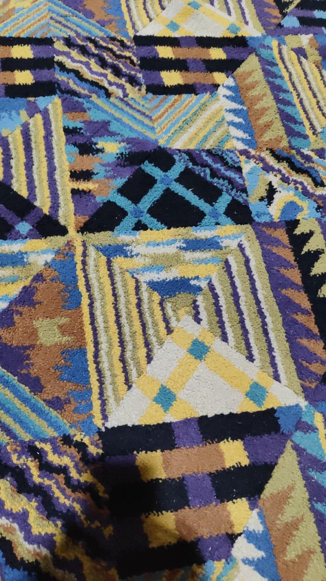 Ce tapis Ottavio Missoni mesure 235 cm.  x  166 cm. est fabriqué en pure laine dans les années 1980. Il présente une combinaison harmonieuse de tons bleus, jaunes et bruns, évoquant une palette vibrante de couleurs riches et de motifs géométriques