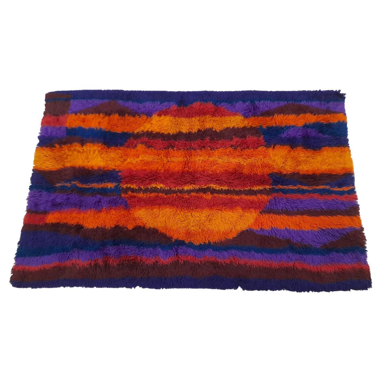 Dieser wunderschöne Rya-Teppich des renommierten dänischen Unternehmens Ege Taepper erinnert mit seinem auffälligen zentralen Muster an einen Sonnenuntergang, bei dem die Sonne in leuchtenden Orangetönen vor einem violetten und blauen Himmel