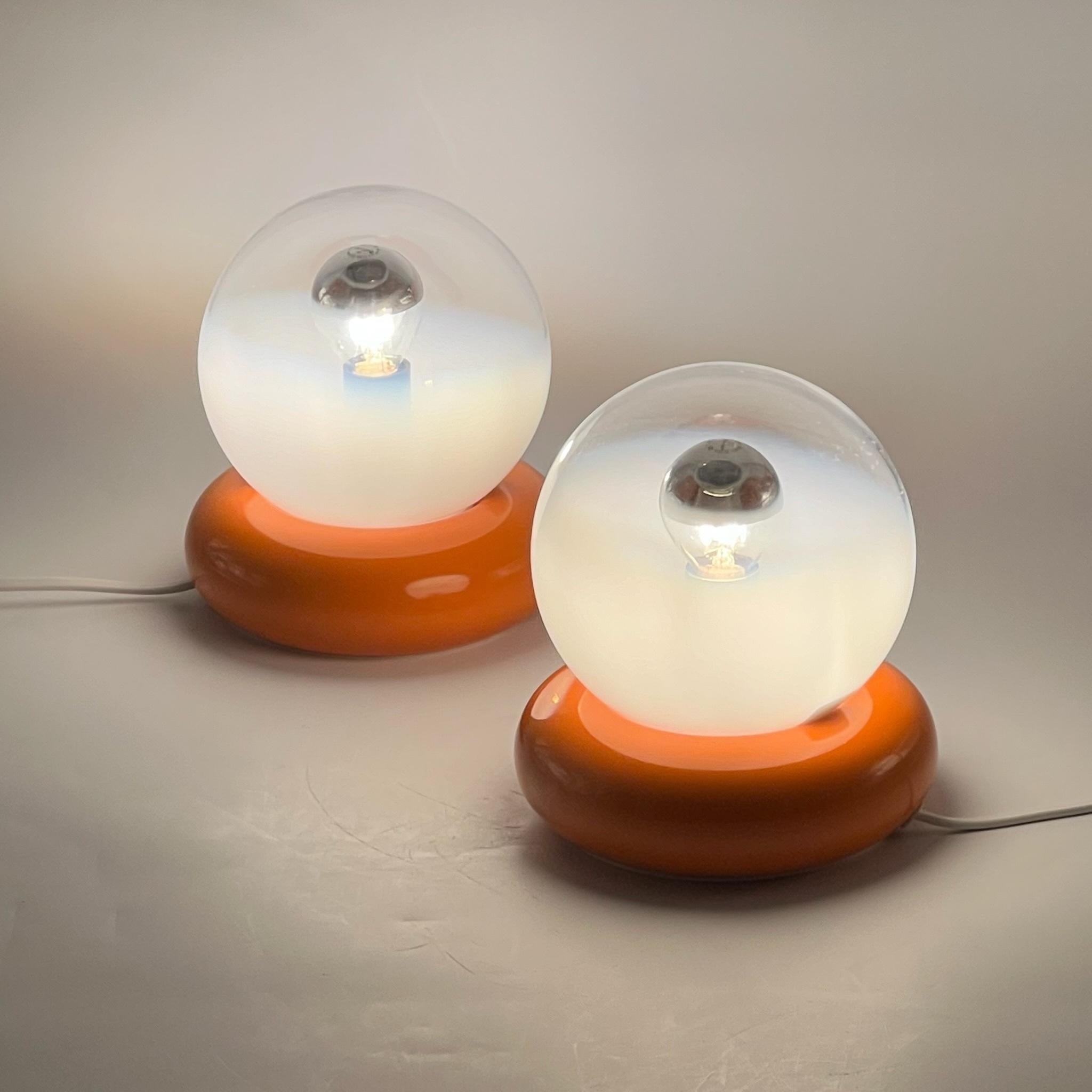 Spruce up your space with this exquisite pair of vintage Targetti Sankey table lamps, showcasing a unique blend of vibrant orange metal bases and handcrafted artistic glass spheres. The glass globes are transparent with subtle white shading,