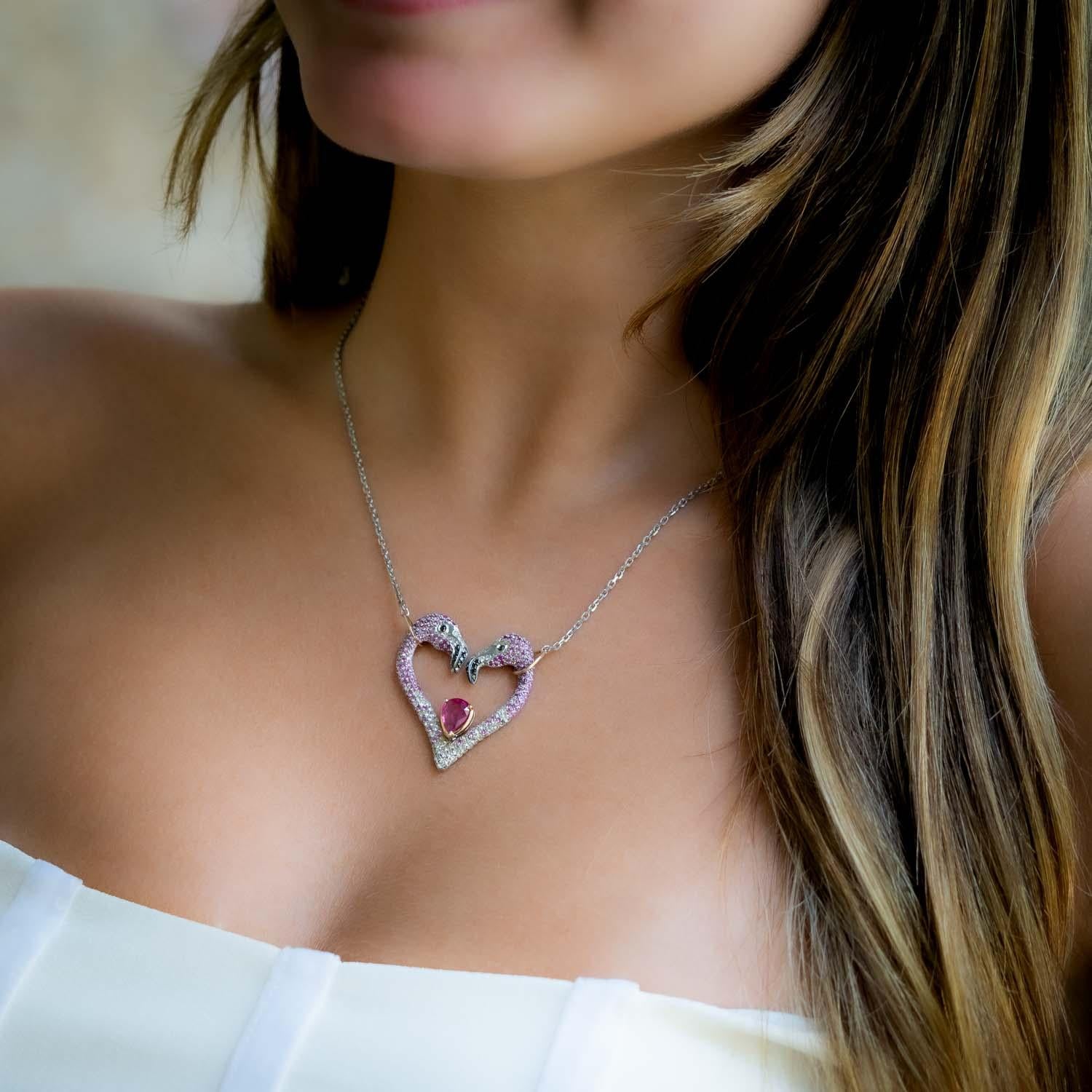 This gorgeous 18 carat rose gold and sterling silver Flamingo Heart Necklace is a unique piece of jewelry. The design features two flamingos forming a heart shape, encrusted with pink and white sapphires. The center stone, a ruby framed in 18K rose