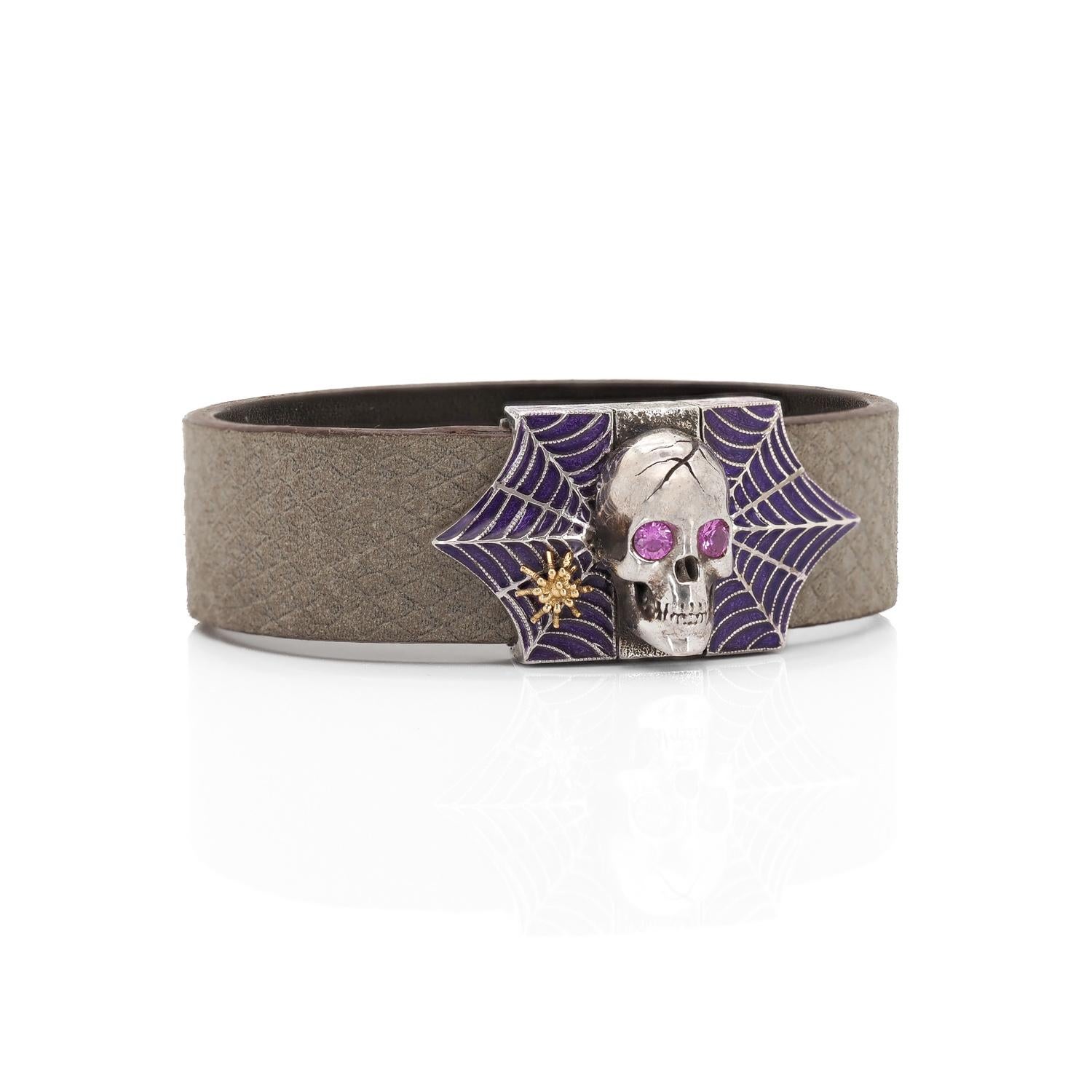 The Skull & Spider Web Bracelet features a 18K gold spider on its silver spider web with purple enamel. The eyes of the skull, set with pink sapphires, adds a special glare to the bracelet. The bracelet is strapped by an embossed grey leather strap,