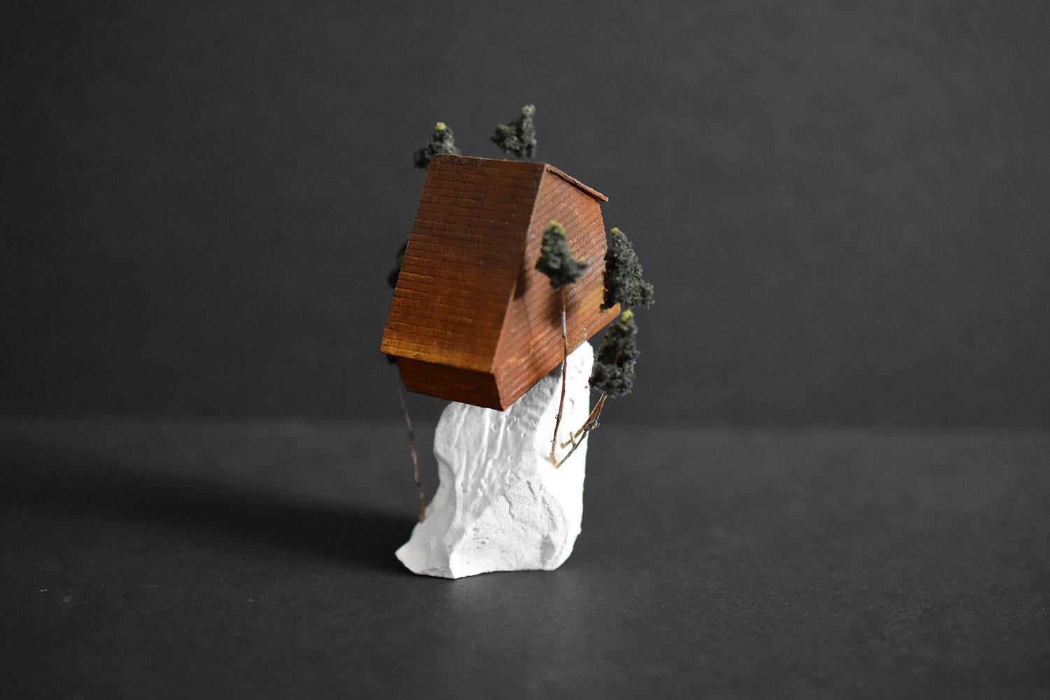 “Between pines, a pause” by Tatiana Flis is a 3.5 x 2 x 2 inch miniature landscape sculpture of reclaimed wood, plastic, and found mixed-media and organic materials. An orange and rust colored salt-box style home sits on top of a white landscape