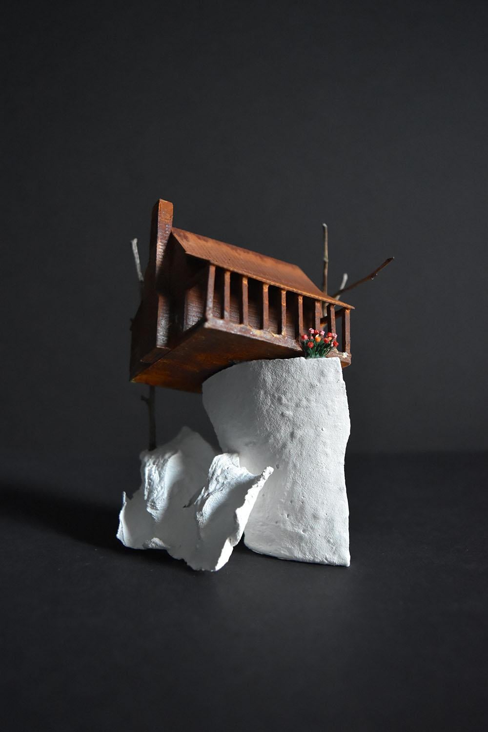 “Everything is Waiting” by Tatiana Flis is a 4.5 x 4 x 4 inch miniature landscape sculpture of reclaimed wood, plastic, and found mixed-media and organic materials. An orange and rust colored building with an exposed porch sits on top of a white