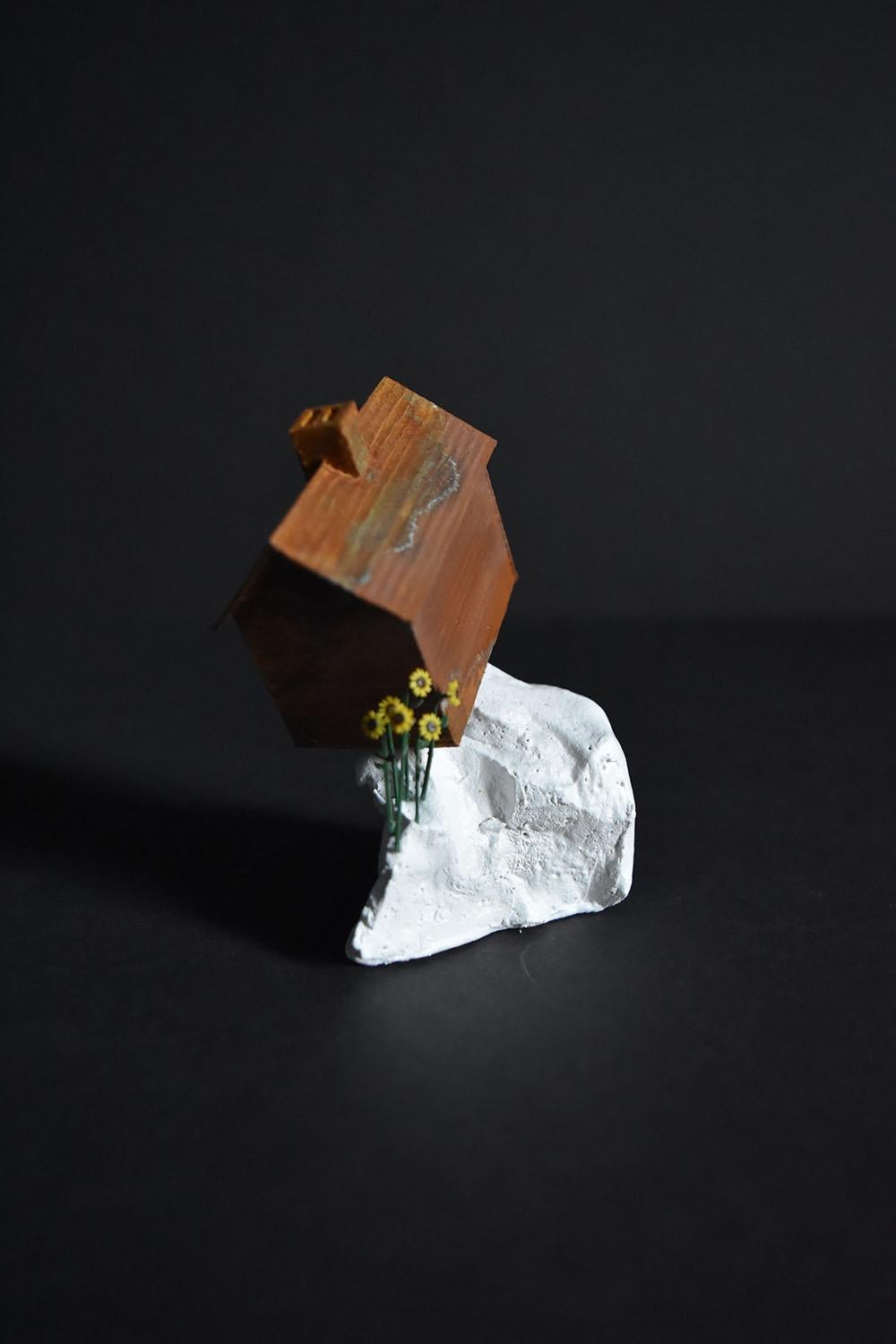“Precarious Ledge” by Tatiana Flis is a 3 x 2 x 2.5 inch miniature landscape sculpture of reclaimed wood, plastic, and mixed-media materials. An orange, rust, and sea foam green patina colored home with a double chimney sits on top of a white empty