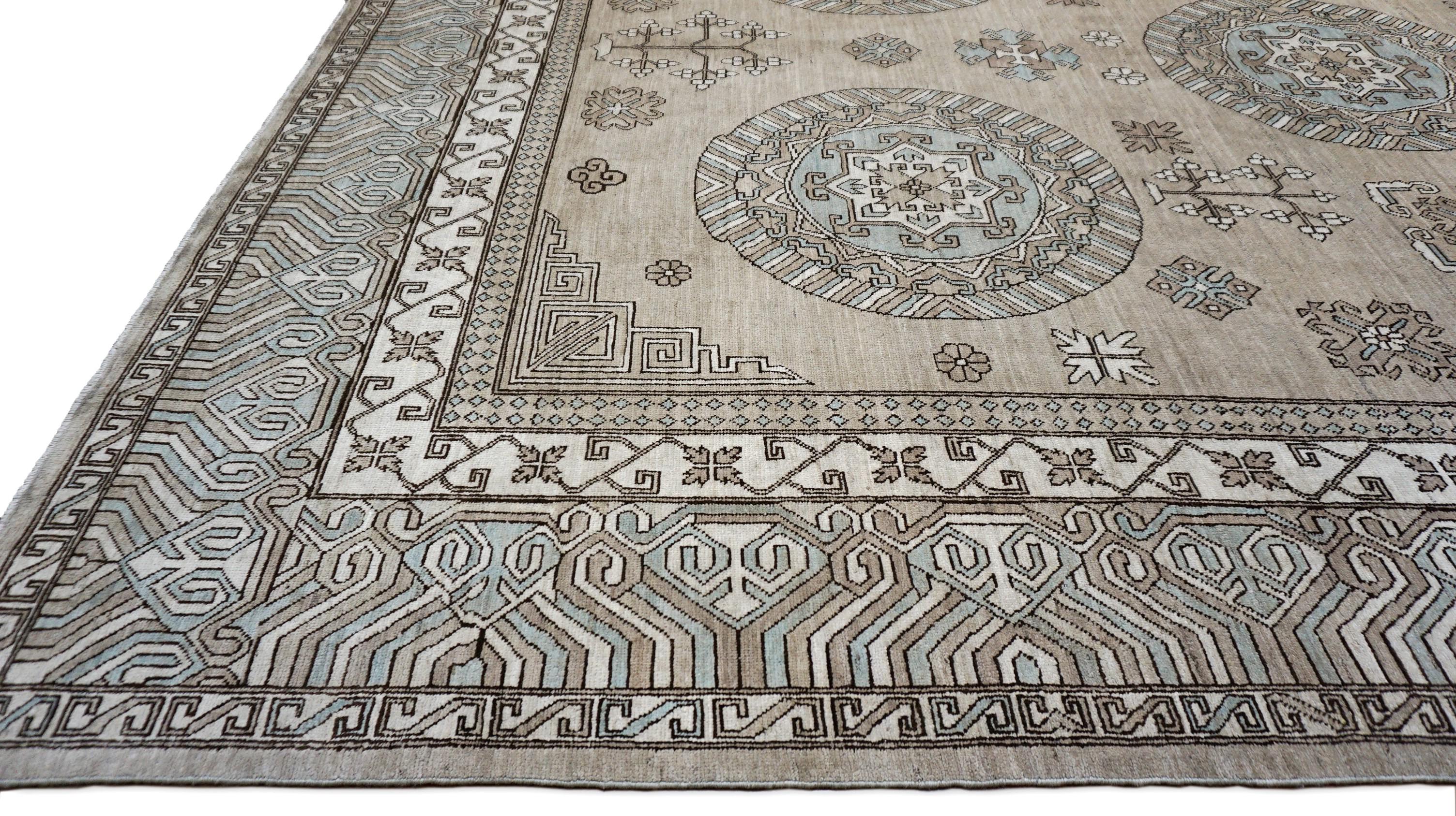 This hand-knotted wool rug is a sophisticated reinterpretation of the classic Khotan design, featuring an elegant blend of geometric medallions, intricate floral motifs, and delicate tribal patterns. Set against a soft tan, taupe, blue, and gray