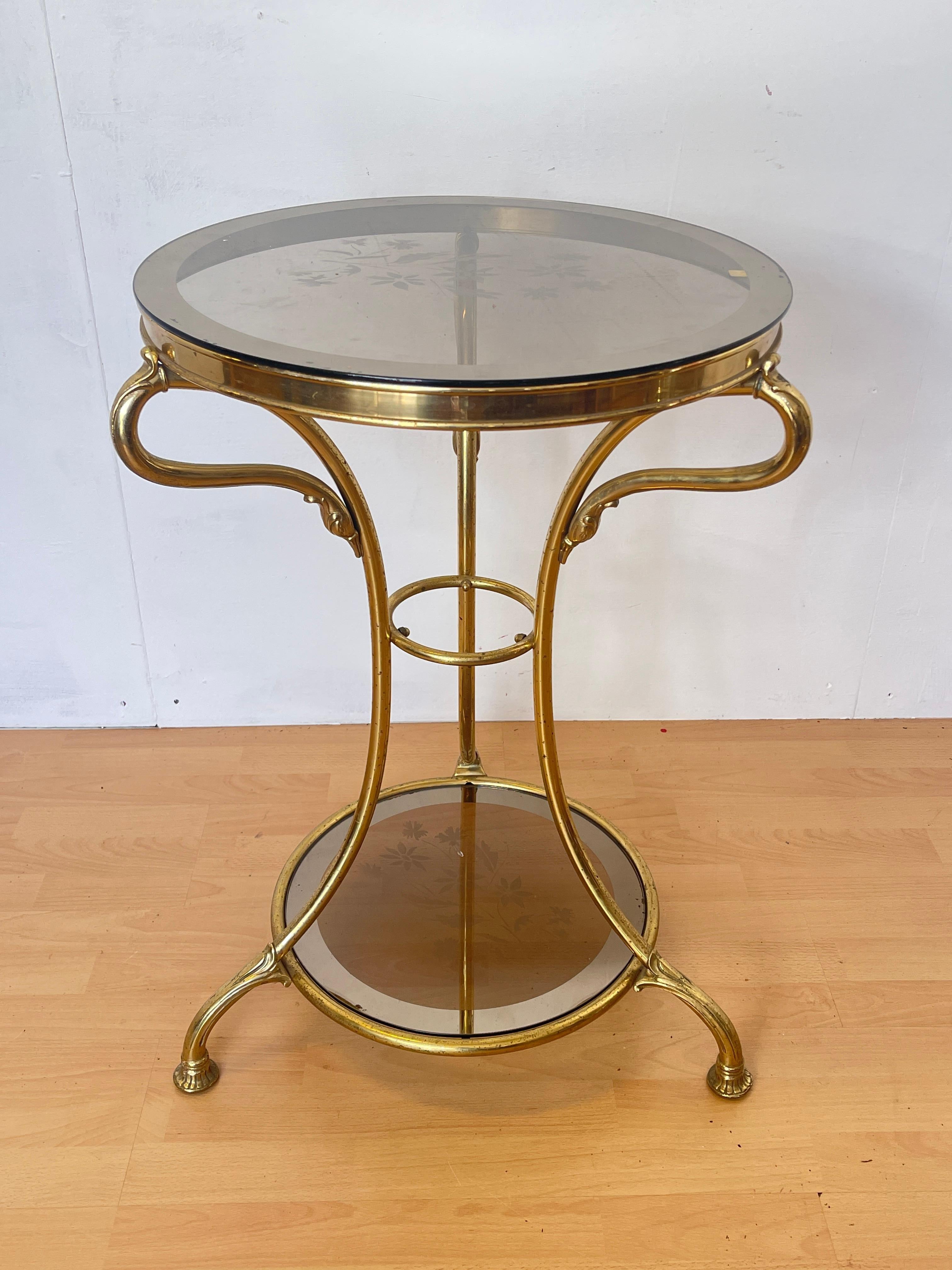 Raffinato tavolino vintage, caratterizzato da una base dorata curvata e un elegante piano in vetro decorato. Il vetro presenta un delicato motivo floreale inciso, creando un gioco di luci e riflessi che arricchisce l’ambiente. Il design sofisticato