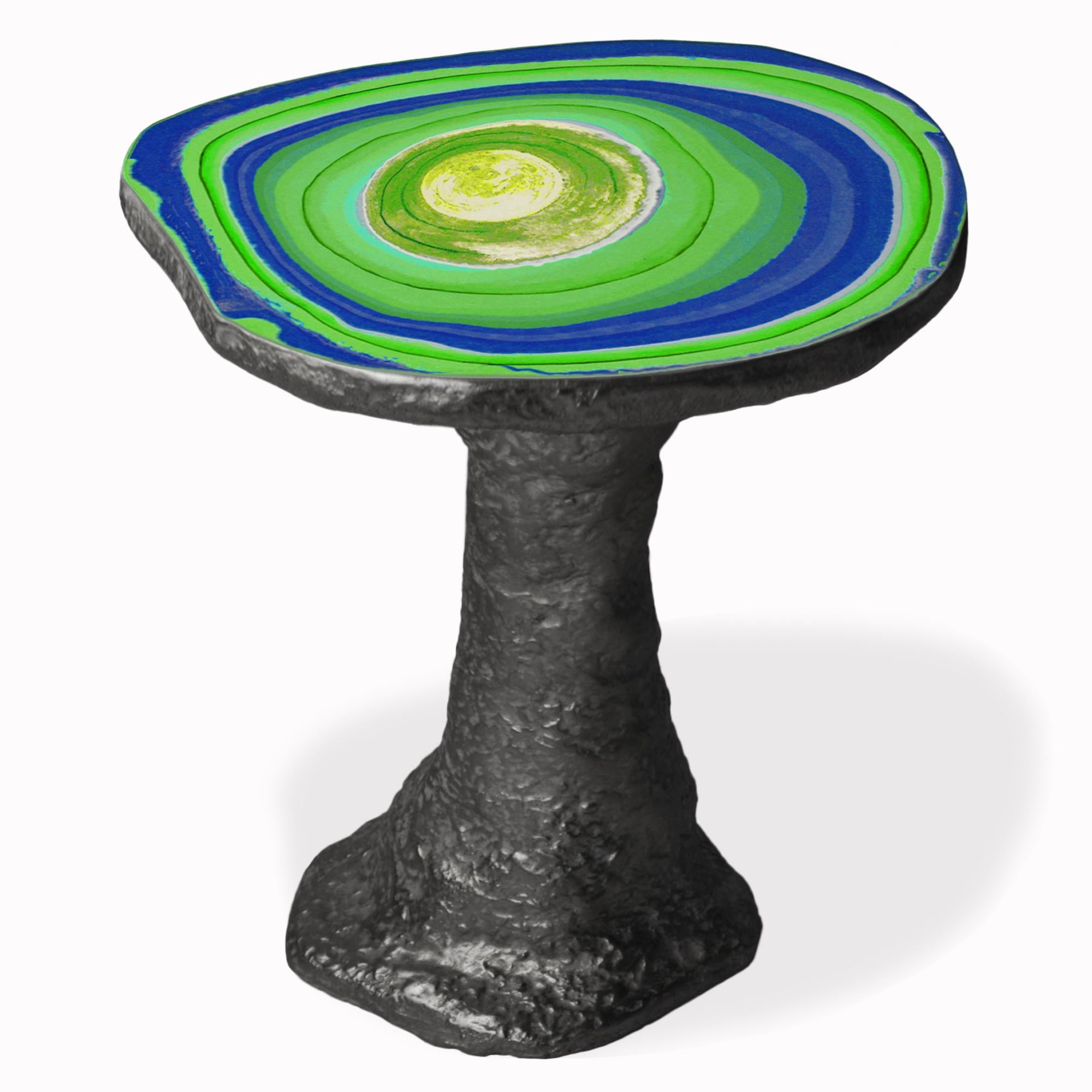 Une table basse réalisée en un seul corps et caractérisée par le contraste entre la surface lisse et polie du plateau en scagliola, aux teintes vertes et bleues audacieuses, et l'aspect rugueux et multiforme de la tige, créant un dialogue entre