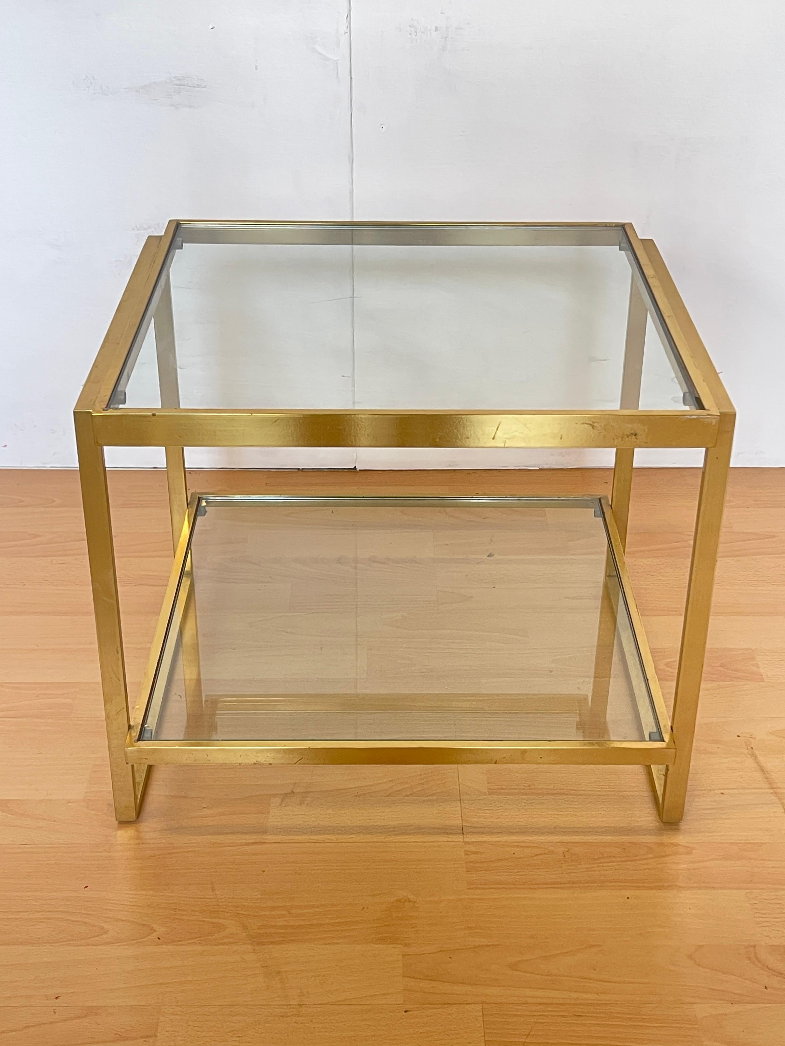 Elégante table basse vintage datant des années 1970, composée d'un cadre en laiton doré et d'une double tablette en verre transparent. Ses lignes épurées et géométriques expriment parfaitement le style moderniste de l'époque, avec des références au