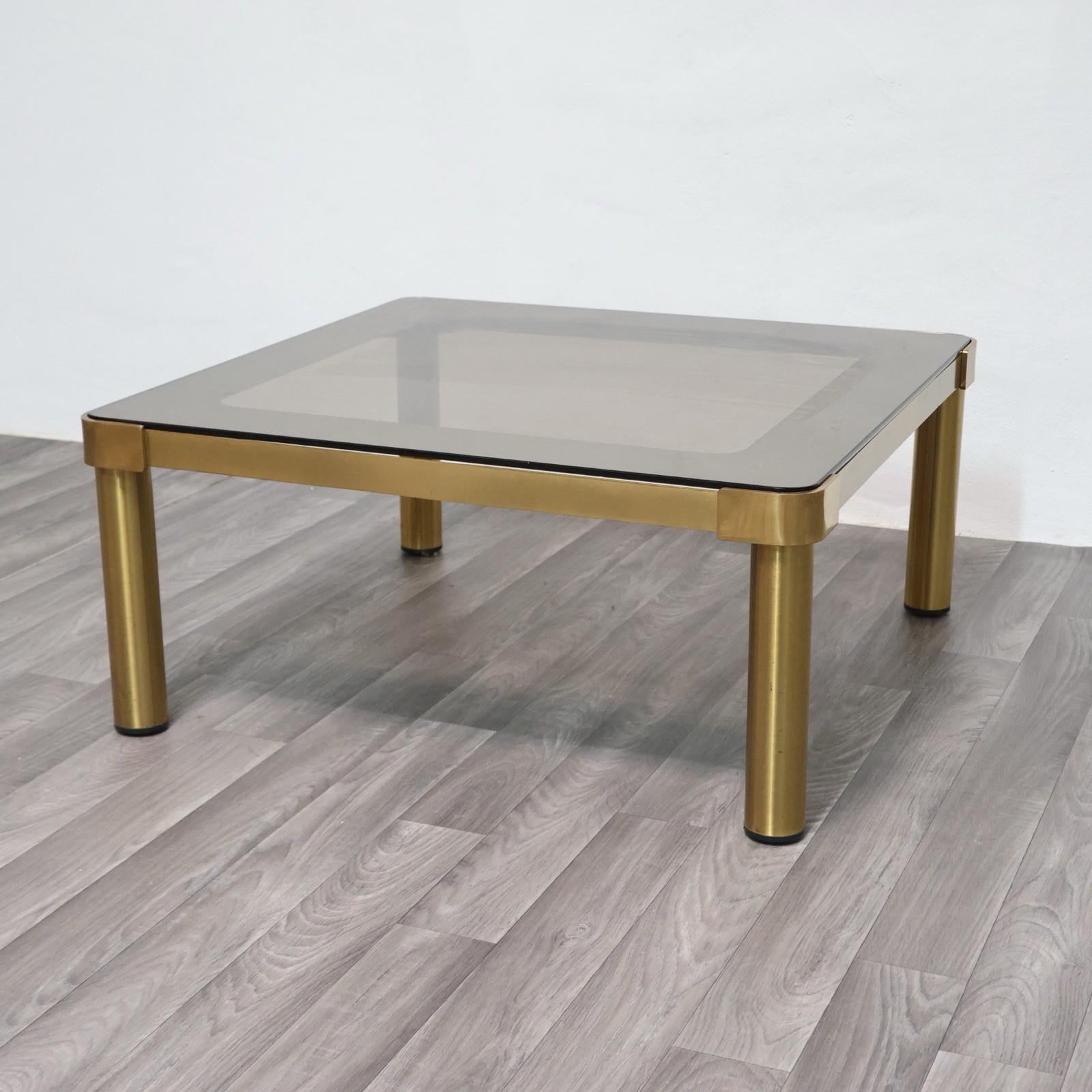 Elégant tavolo basso da salotto in perfetto stile Hollywood Regency, databile agli '70. Le meuble est en ottone dorato lucido, avec des joues cilindriche robustes et des détails angolari smussati, tandis que le piano est en vetro fumé, spesso et